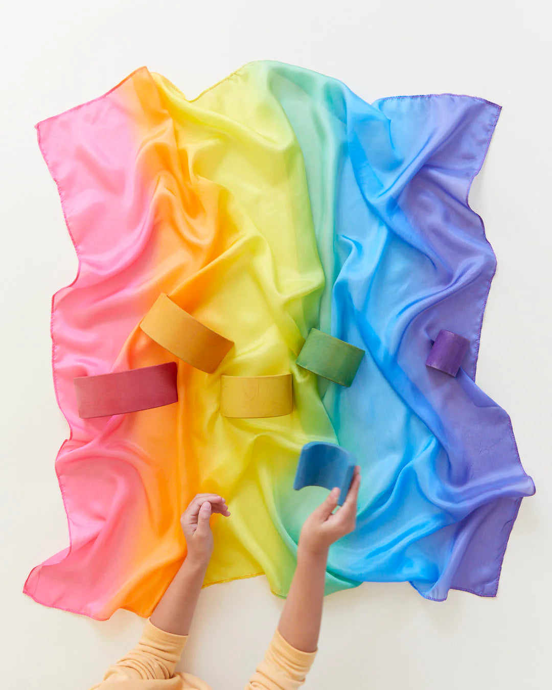 An Enchanted Playsilk Rainbow from SARAH'S SILKS, crafted entirely of 100% pure silk, is draped across a white surface with multiple cardboard tubes scattered on top. Two hands reach for the tubes, with one grasping a blue tube. The playsilk displays a beautiful transition of gradient colors from pink to purple, encouraging imaginative and open-ended play.