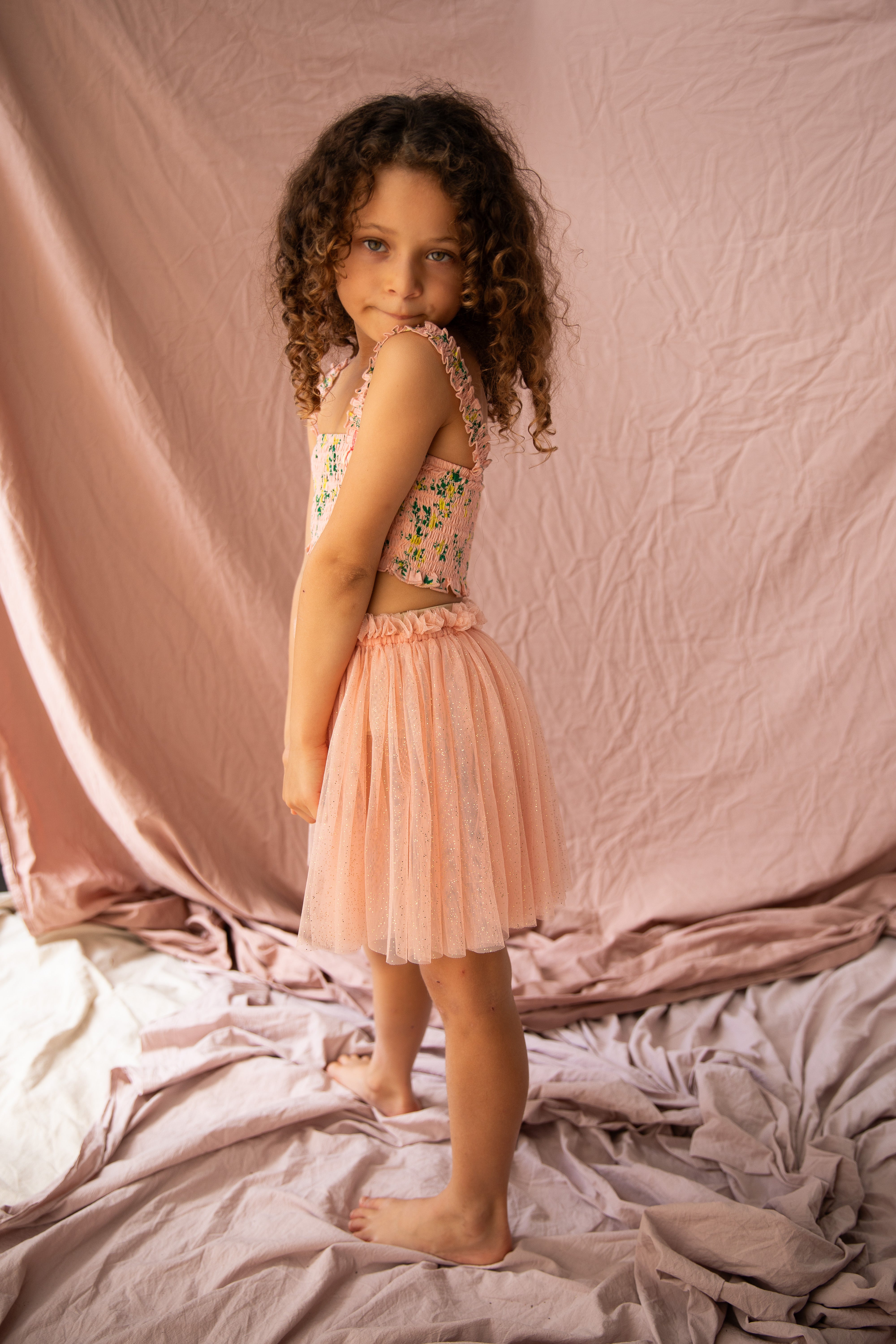 A young girl with curly hair stands on crumpled pink fabric, wearing a floral top and a Classic Tutu Pink Bloom from BELLA + LACE. The background is a matching soft pink fabric, creating an elegant and delicate setting.