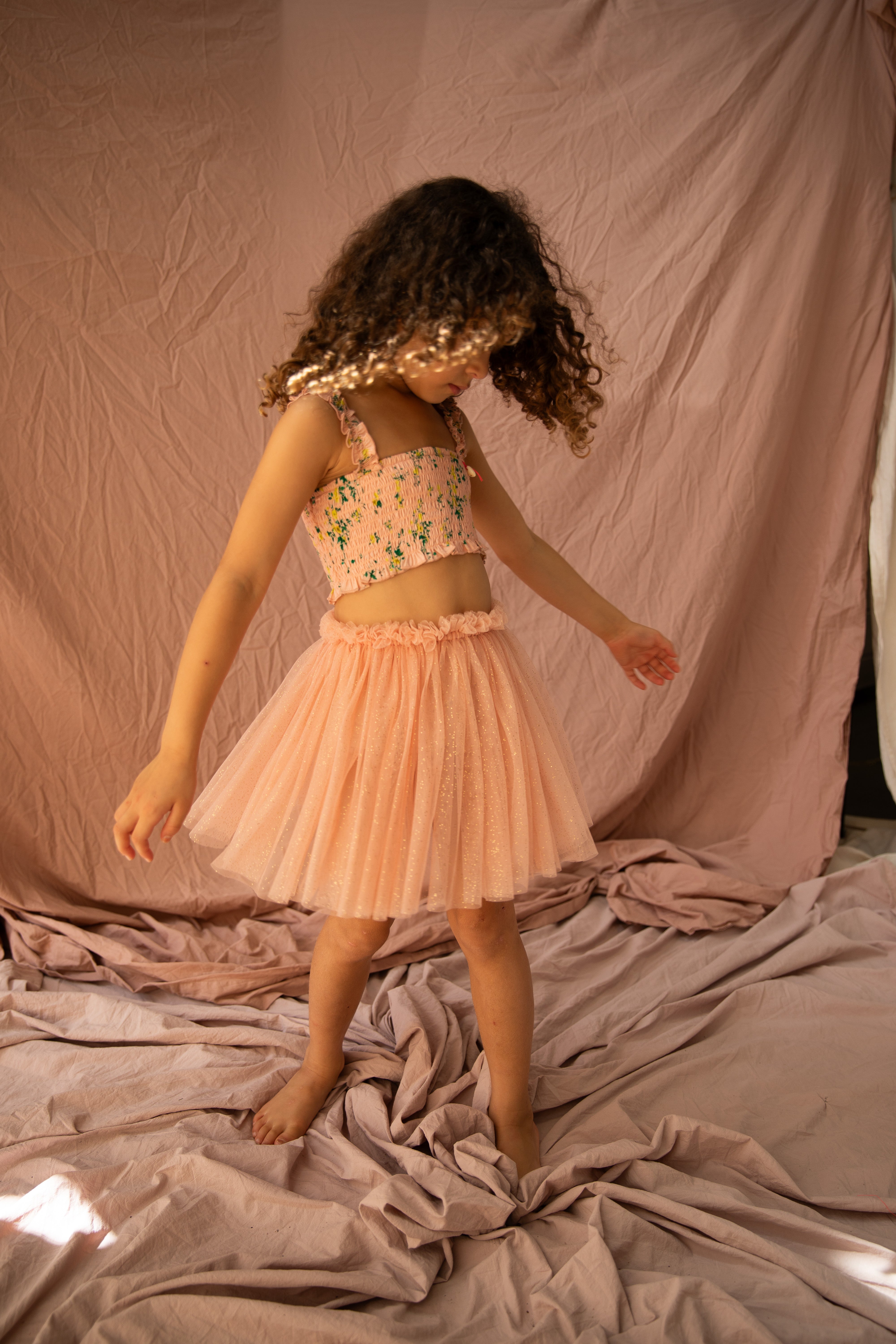 A young girl with curly hair, wearing the BELLA + LACE Classic Tutu Pink Bloom, dances barefoot on a soft pink fabric backdrop.