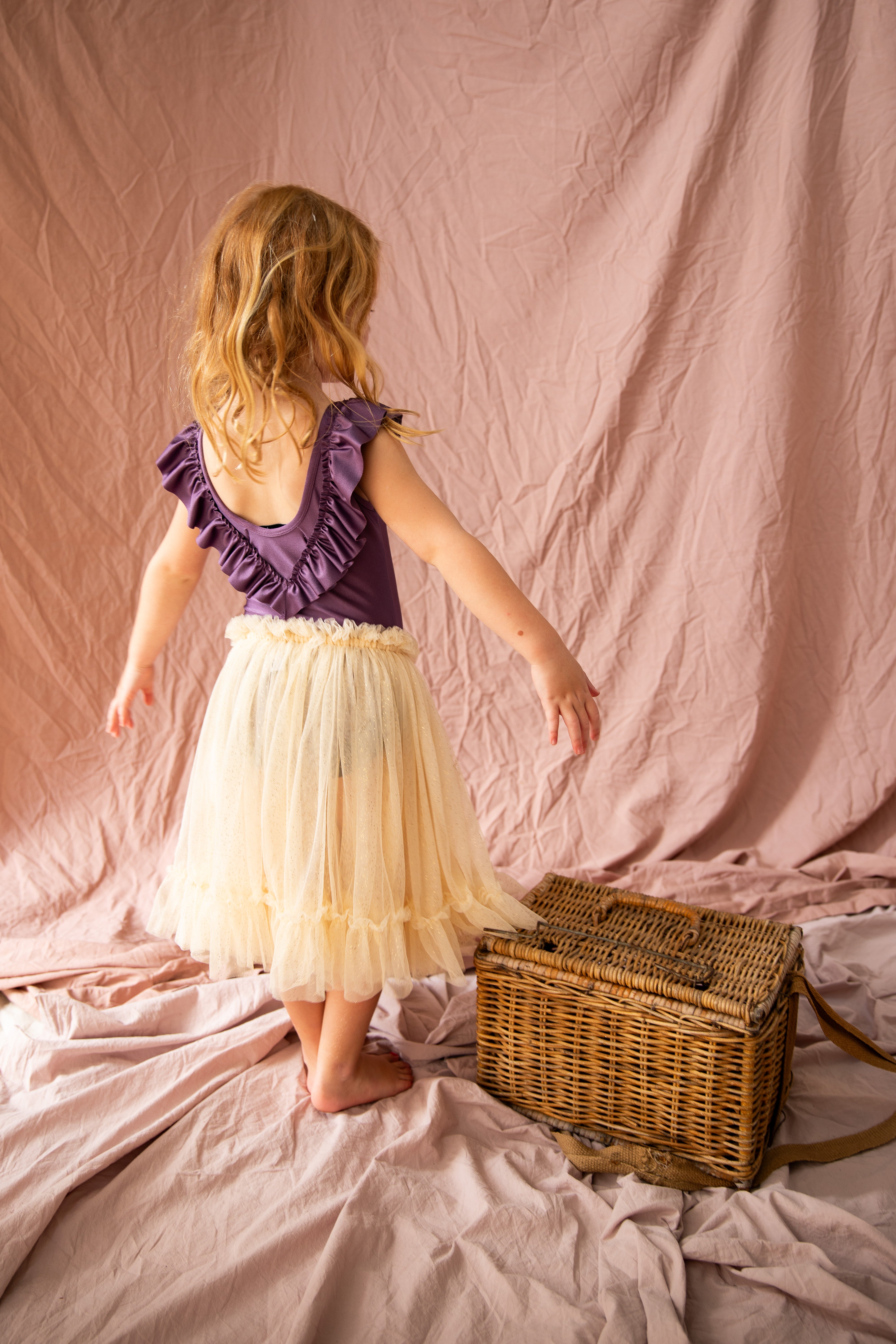 A young girl with blonde hair stands on a pink fabric backdrop, wearing a BELLA + LACE purple top with ruffled sleeves and a Carrie Tutu Vanilla. She is barefoot and facing away, with one hand on a wicker basket.