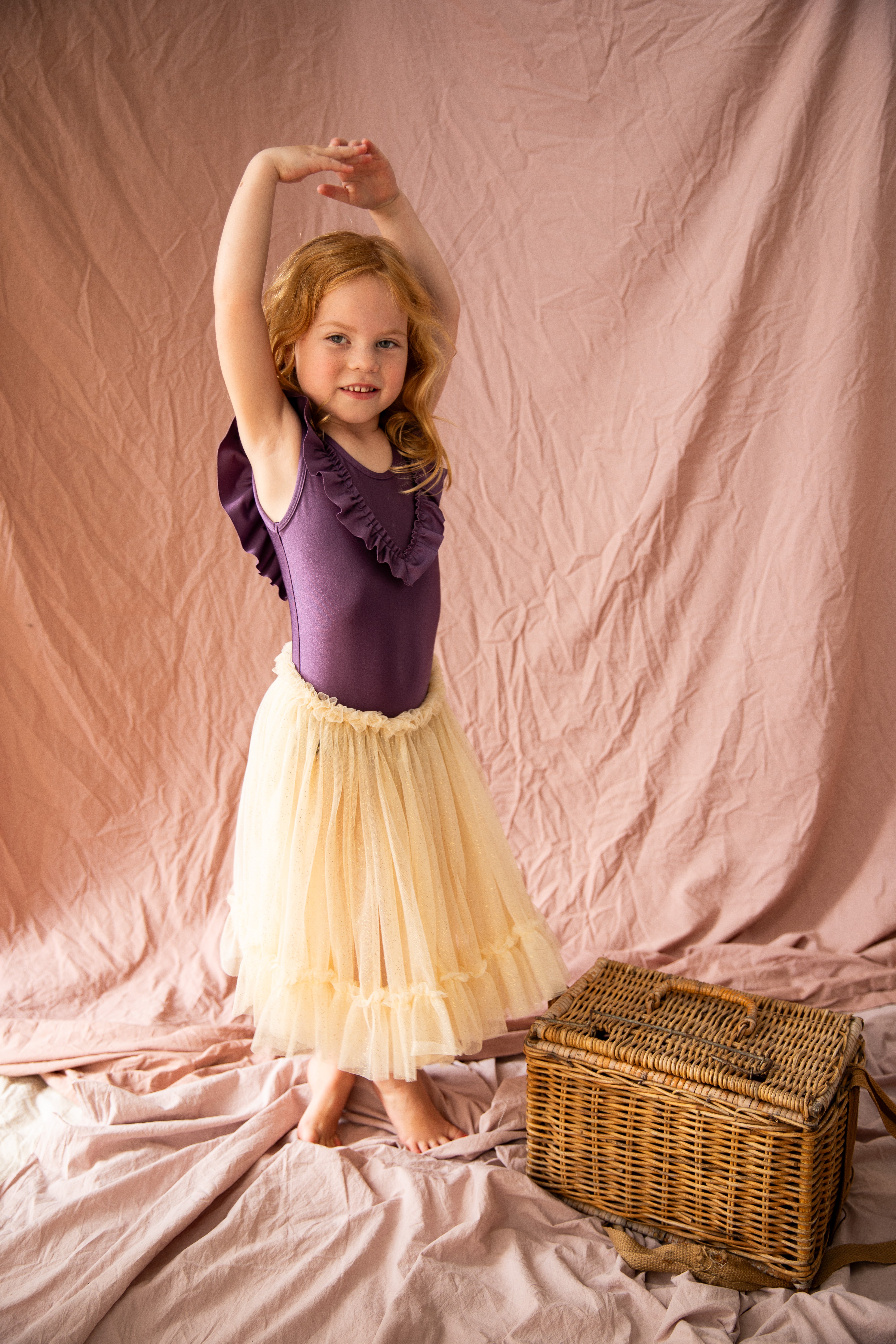 A young girl with red hair poses gracefully in the Carrie Tutu Vanilla—a classic ensemble by BELLA + LACE, featuring a purple leotard paired with a cream tulle skirt. She stands on a pink fabric backdrop next to a wicker basket, her arms elegantly raised above her head.