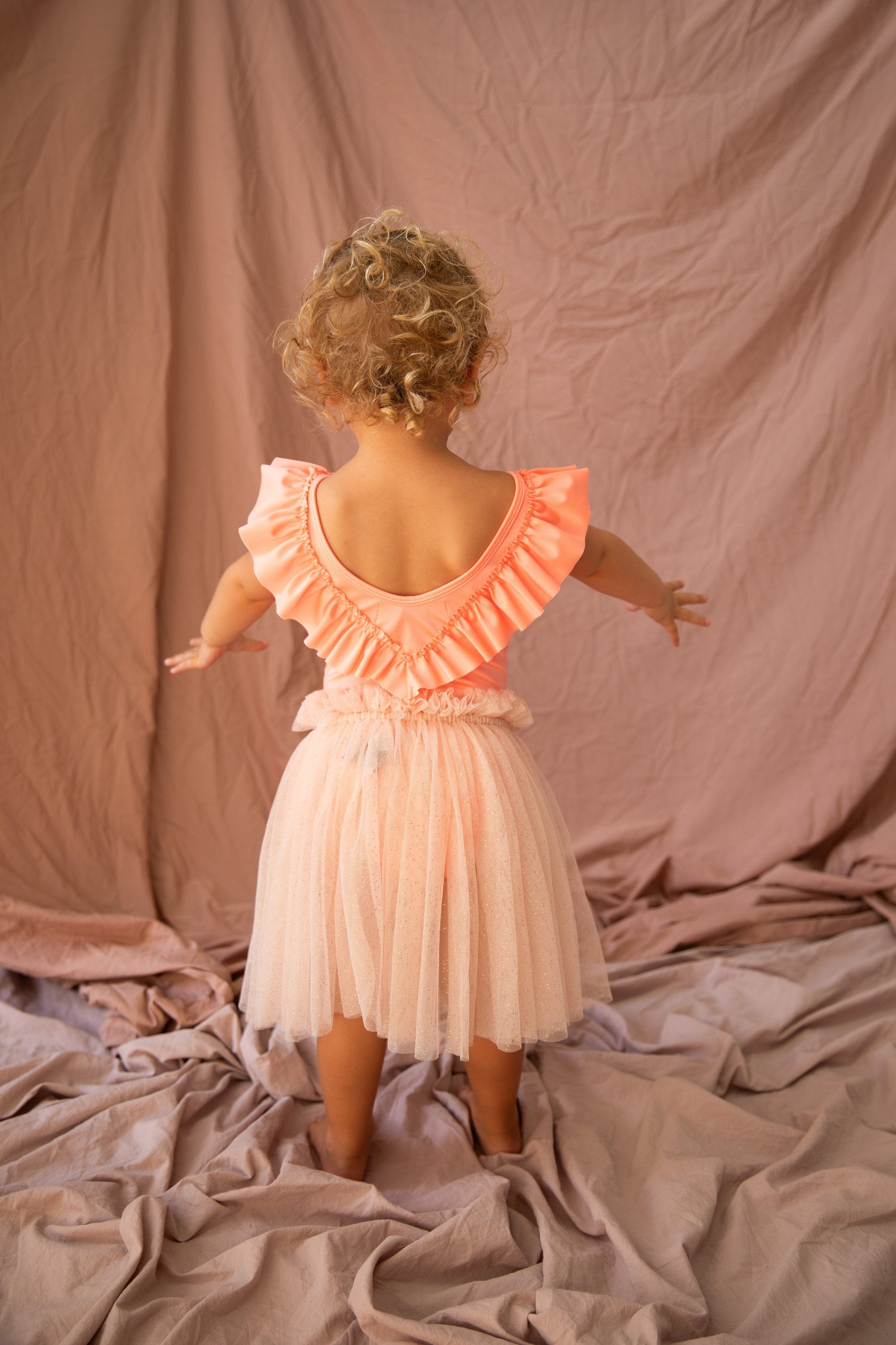 A young child with curly blond hair is standing with their back to the camera, adorned in the Roxette One Piece Prawn by BELLA + LACE. The peach-colored dress features ruffled sleeves and a tutu-style skirt, making it an ideal addition to any summer wardrobe. The soft, light brown fabric draped in the background enhances the elegance of the scene.