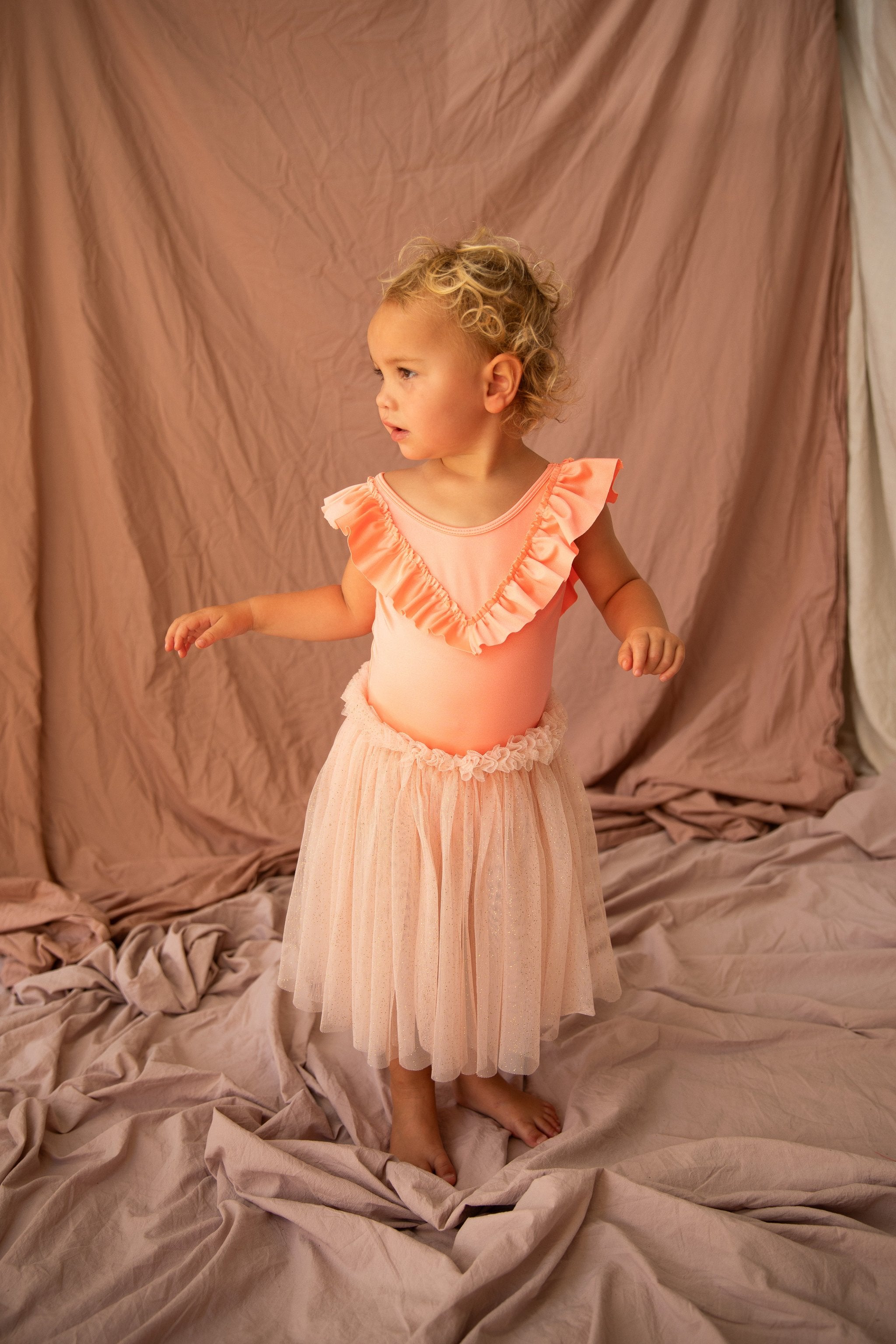 A toddler with curly blonde hair stands on wrinkled pink fabric, dressed in the Roxette One Piece Prawn by BELLA + LACE. This peach-colored outfit, featuring a ruffled top and a tulle skirt, adds a touch of whimsy to their summer wardrobe. The child appears to be looking attentively to the side against the matching background.