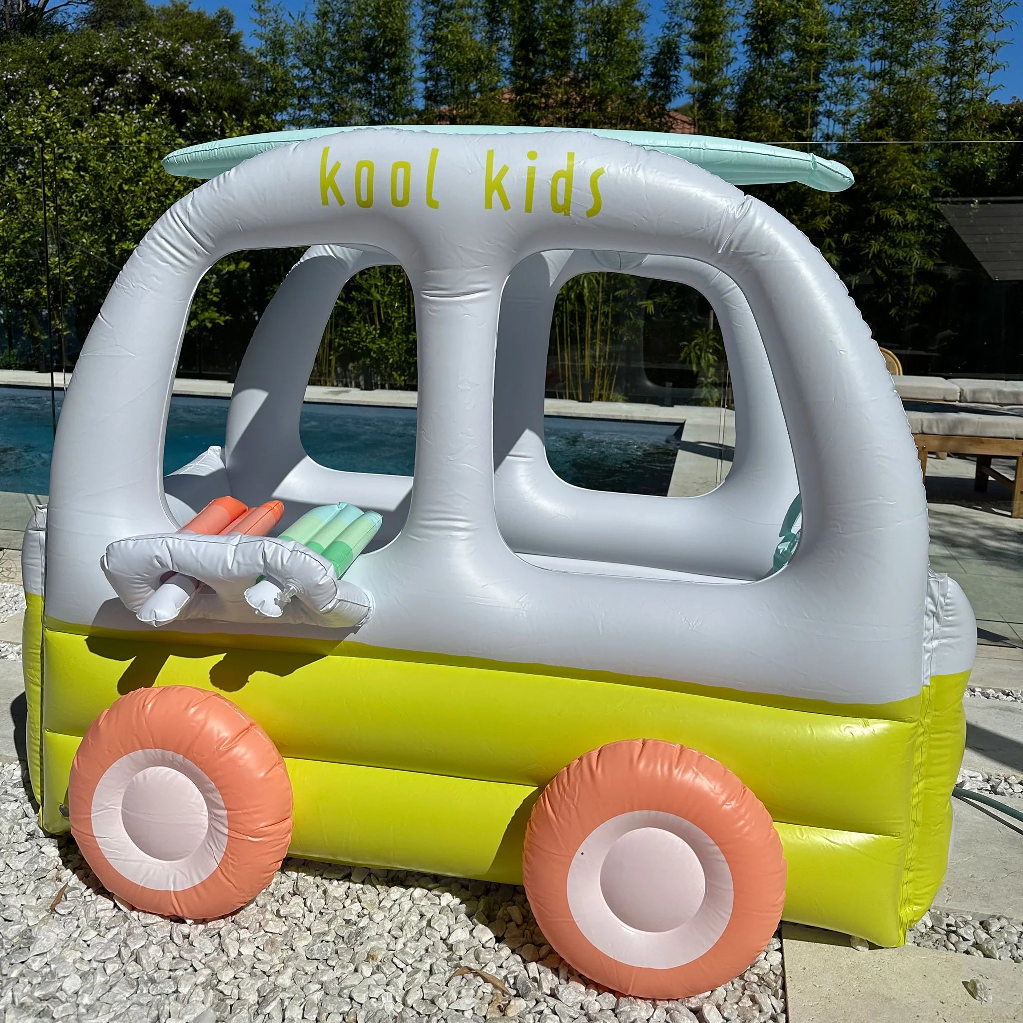 The Inflatable Cubby Ice Cream Van by SUNNYLIFE features pastel-colored details, including pink wheels and a surfboard on top labeled "kool kids." Made from durable PVC that's non-toxic and Phthalate free, this inflatable pool toy is set near a swimming pool with trees and a fence in the background.