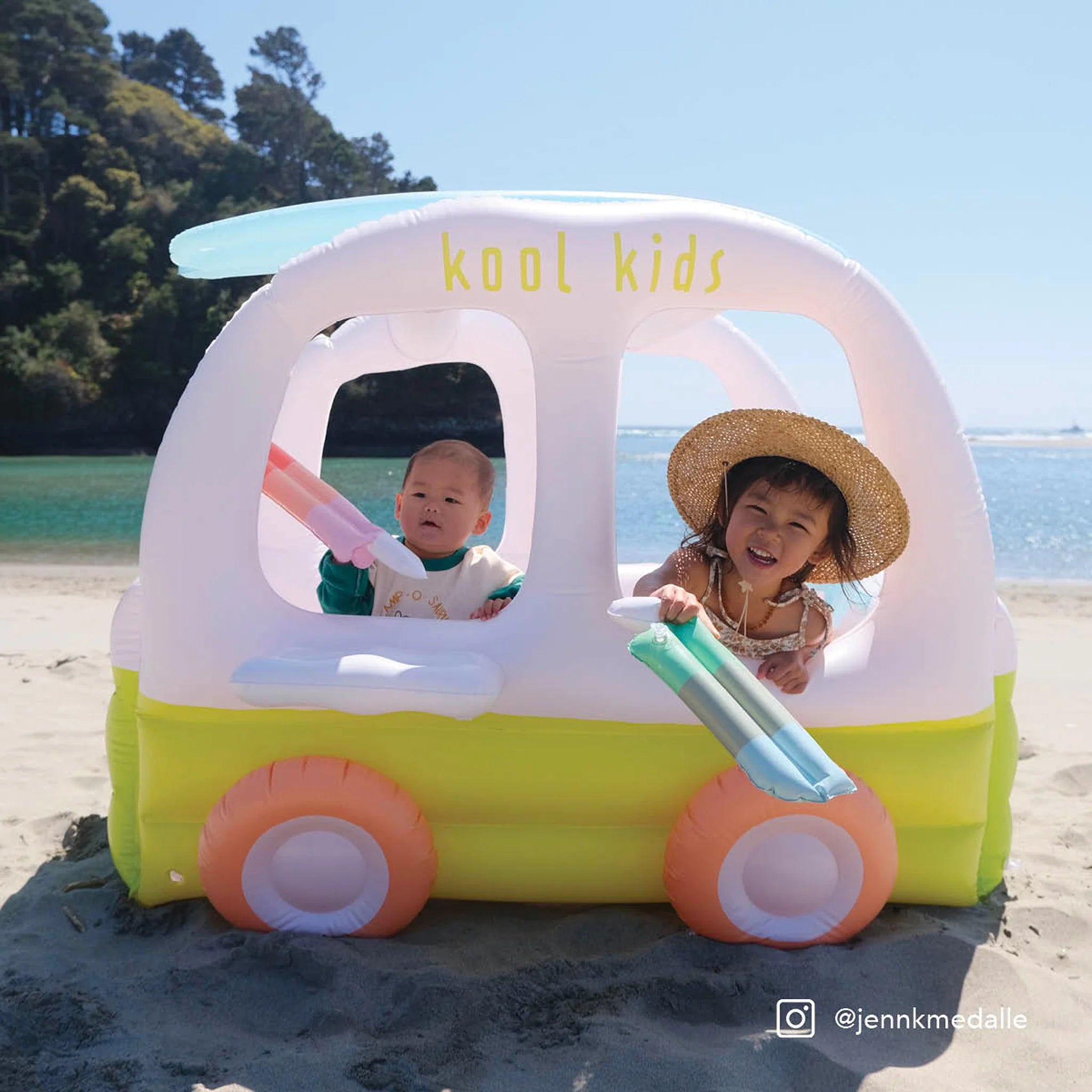 Two children are playing in a SUNNYLIFE inflatable toy called the "Inflatable Cubby Ice Cream Van" on a sunny beach. One child is wearing a white outfit, and the other is sporting a sun hat, smiling. The inflatable, made of durable PVC and non-toxic phthalate-free materials, floats on clear blue water with trees in the background.