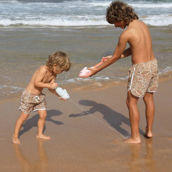 Two children are playing on a sandy beach near the ocean, both wearing matching swim trunks. One child uses a SUNNYLIFE Shark Water Squirter, showcasing its maximum soaking capability by squirting water at the other child, who is laughing and holding a similar toy. Waves ripple in the background.