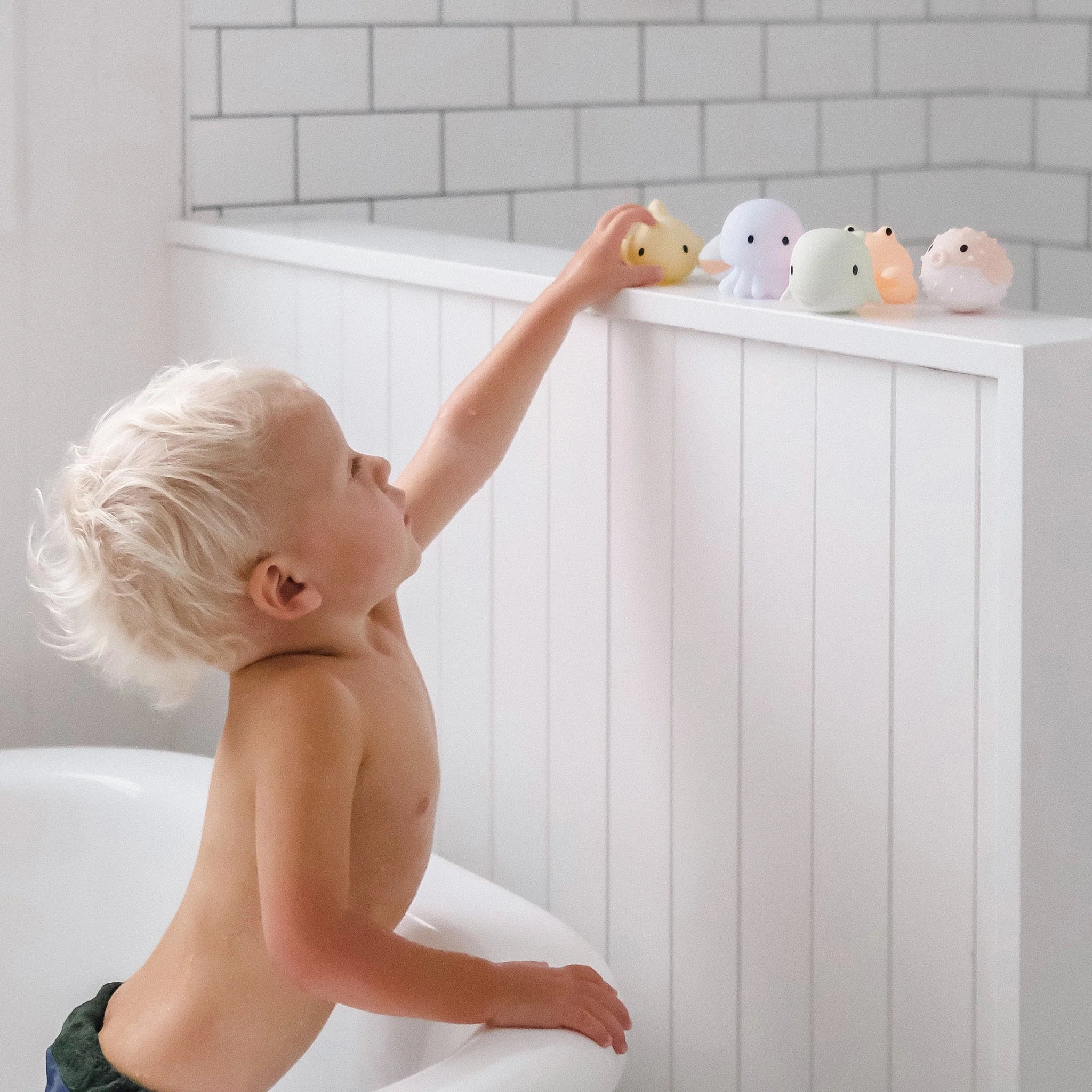 A young child with blonde hair reaches up to place a yellow Sea Animal Splash Toy by SUNNYLIFE, which floats on water, on the edge of a white bathtub. Other colorful splash toys, including an octopus, are already lined up for play. The bathroom has white paneling and white subway tile walls.