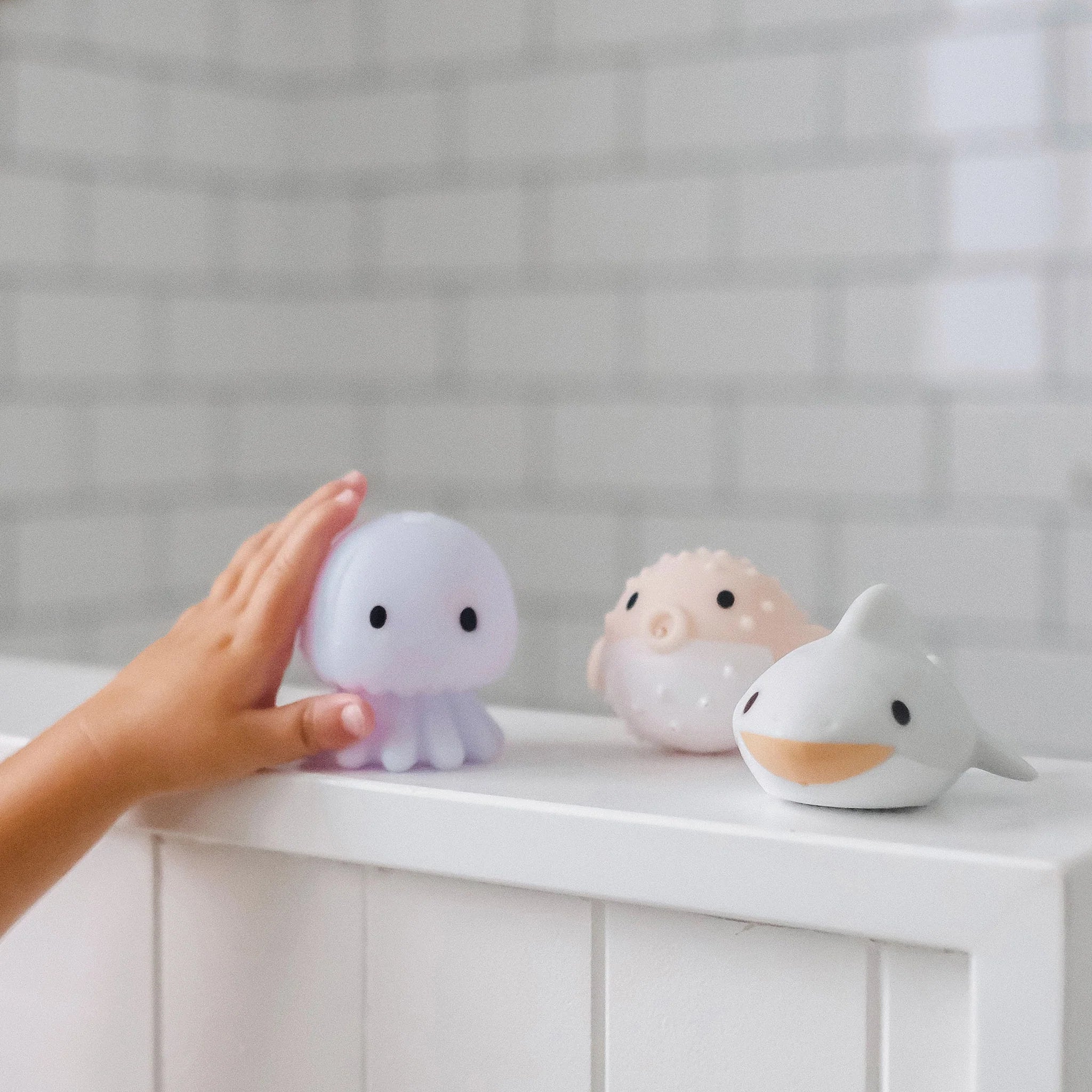 A child's hand reaching towards three Sea Animal Splash Toys by SUNNYLIFE, lined up on a white surface. The light purple octopus, beige puffed fish, and gray shark—each with a cute, smiling face—are perfect for splash play. These fun squirters float on water, adding extra excitement to bath time against the white tiled walls.