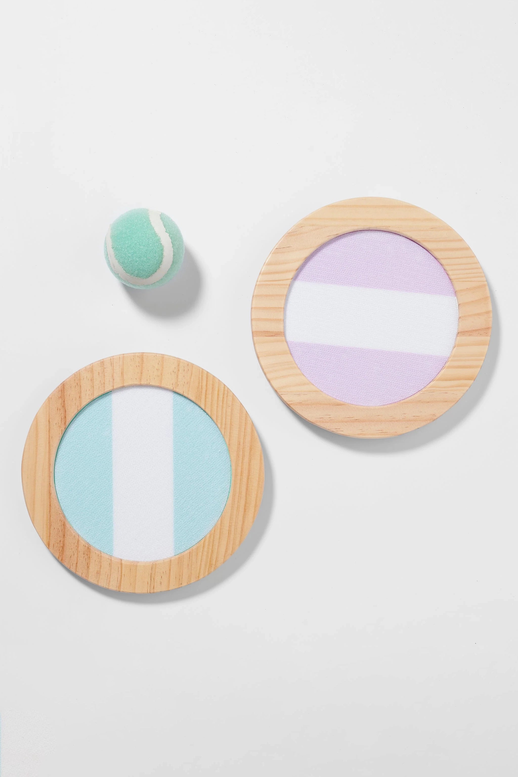 A green tennis ball and two Velcro catching bats from the SUNNYLIFE Catch Ball Set, featuring wooden frames and pastel-colored surfaces, are placed on a white background. One bat boasts a teal and white design while the other showcases lavender and white. An adjustable elastic strap ensures secure gameplay.