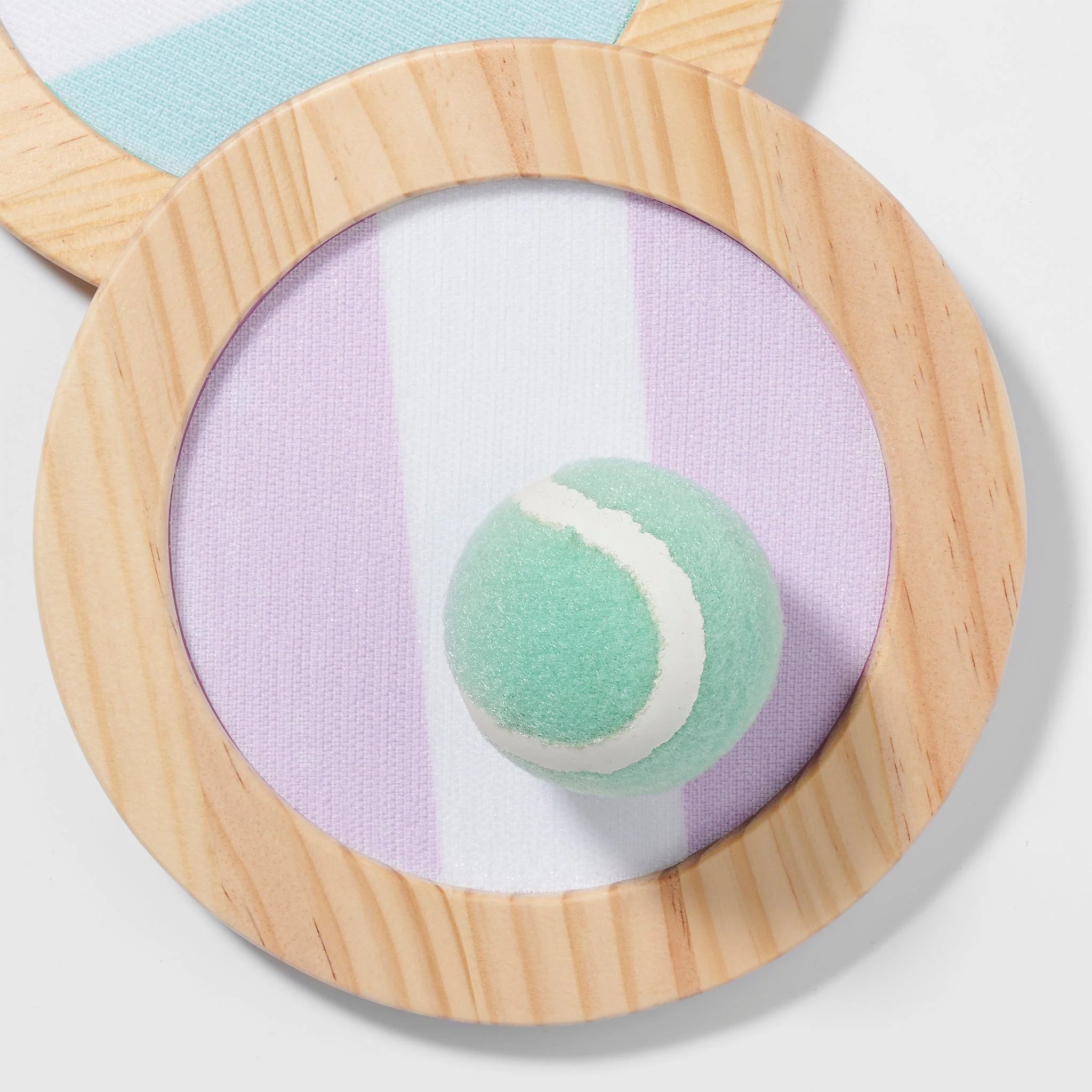 A close-up of SUNNYLIFE's Catch Ball Set reveals a circular wooden paddle with a purple, white, and blue striped surface, holding a green tennis ball with a white stripe. The paddle features an adjustable elastic strap for comfort, and the set includes Velcro catching bats.