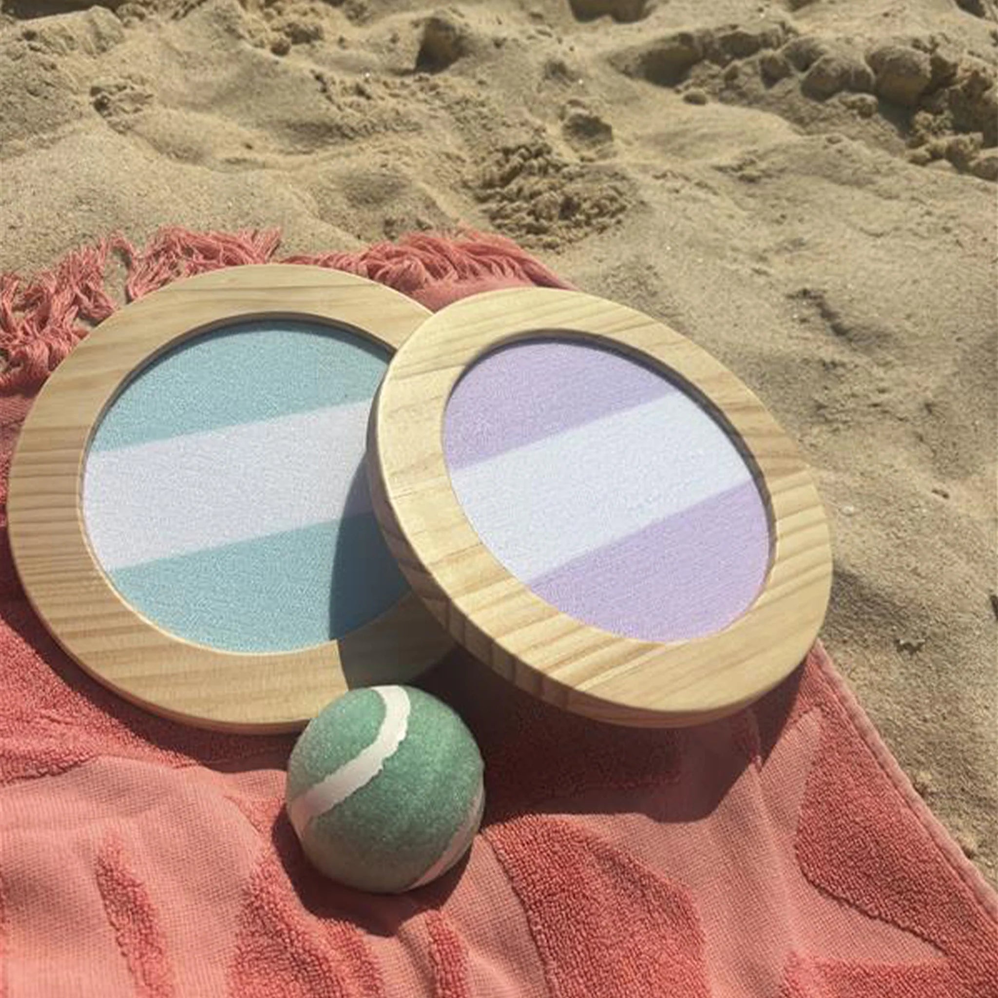 Two wooden-framed round paddles, one with blue and white stripes and the other with purple and white stripes, lie on a peach towel on sandy ground. Nearby, a green and white ball from the SUNNYLIFE Catch Ball Set awaits a game of Velcro catching bats or to be neatly packed away in their reusable carry bag.