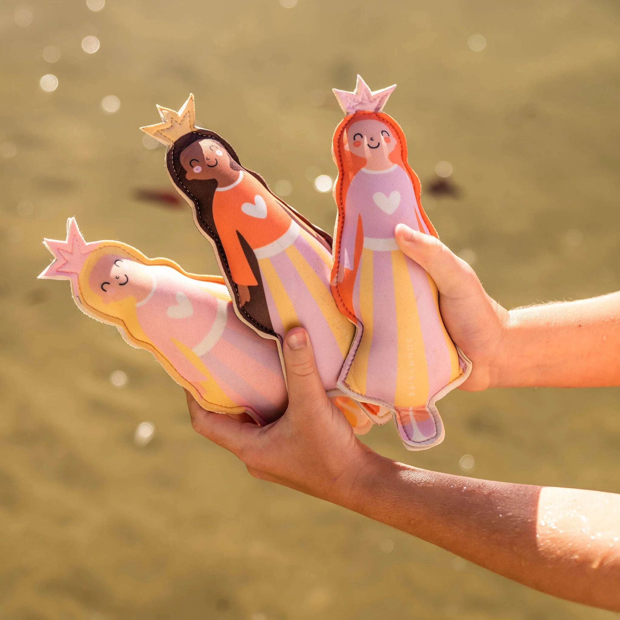 Close-up of two hands holding the Princess Swan Dive Buddies (Set of 3) from SUNNYLIFE. These dolls, made from durable neoprene material, feature simplistic and cheerful faces and are dressed in vibrant skirts with heart motifs on their chests. The slightly out-of-focus background suggests an outdoor setting.