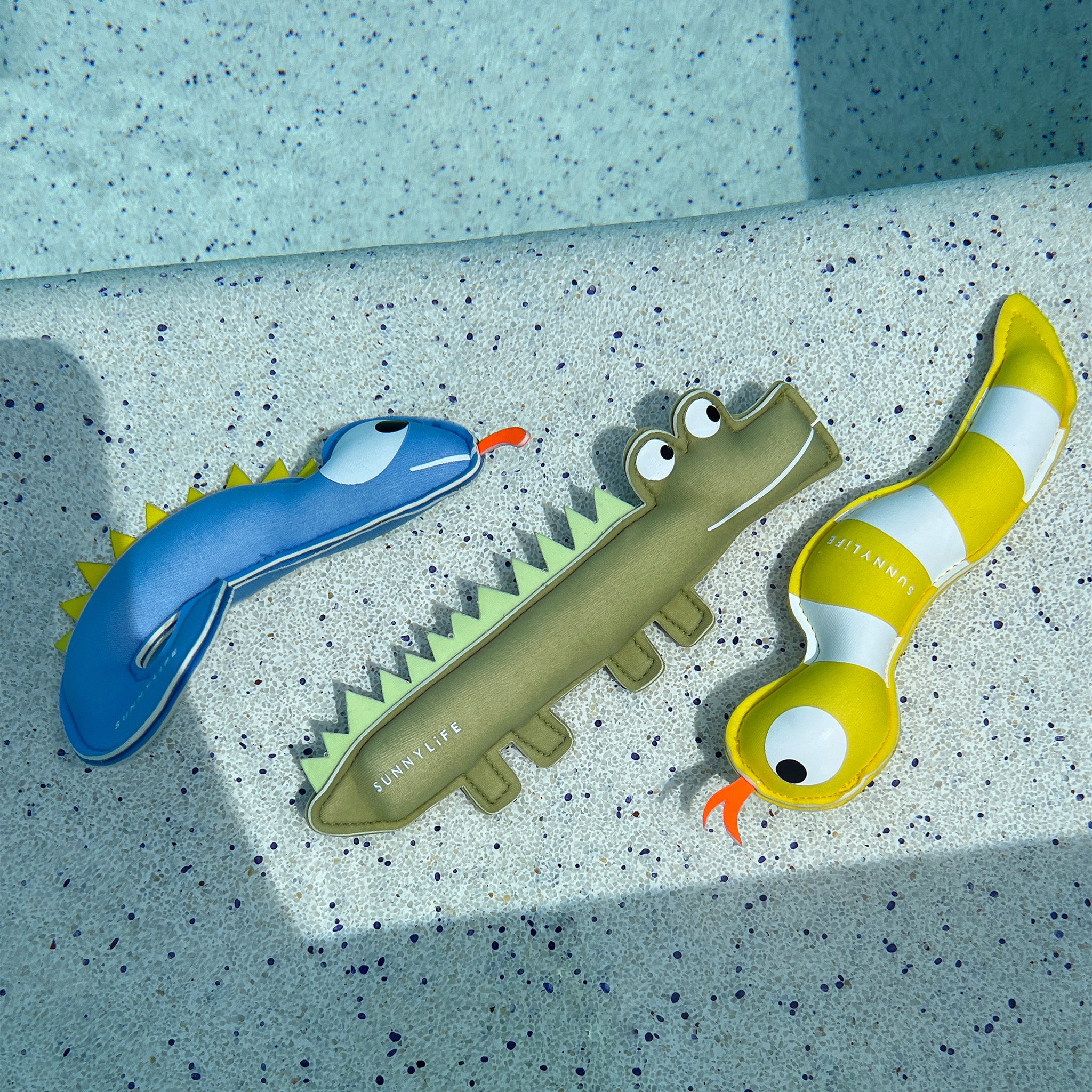 Three inflatable pool toys shaped like animals are arranged on a light-colored speckled surface. This set of 3, named "Into the Wild Dive Buddies" from the brand SUNNYLIFE, includes a blue dinosaur, a green crocodile, and a yellow striped snake. These toys are designed to enhance children's diving skills and feature playful and colorful designs made from durable neoprene material.