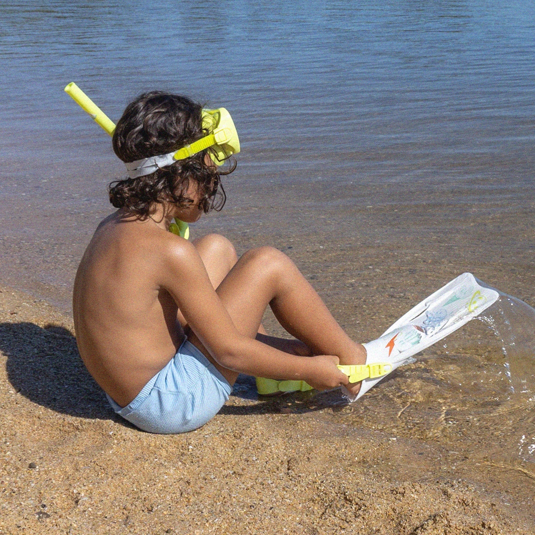 A child with long dark hair is sitting on a sandy beach, wearing the SUNNYLIFE Kids Snorkel Set in Yellow and adjusting one of their flippers near the edge of the water on a sunny day. The calm, blue water is visible in the background.