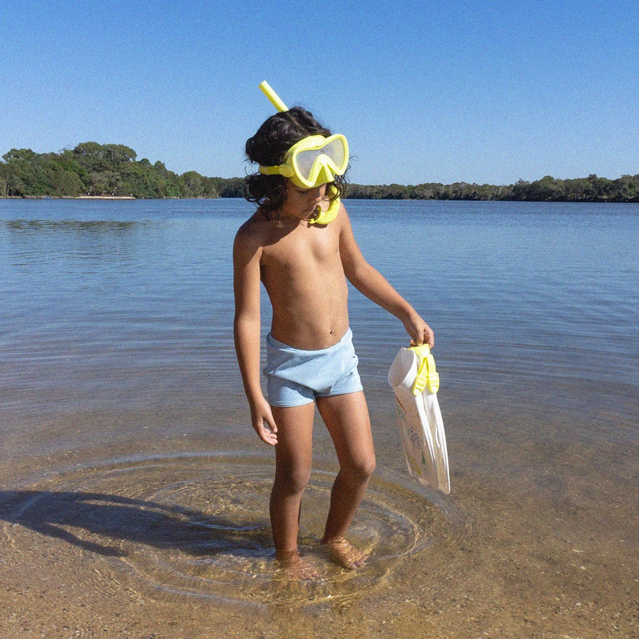 A young child stands in clear, shallow water at a beach, wearing blue swim trunks and the SUNNYLIFE Kids Snorkel Set in Yellow. The child is holding a small white tote bag and gazing down at the water, with a clear blue sky and green treeline in the background.