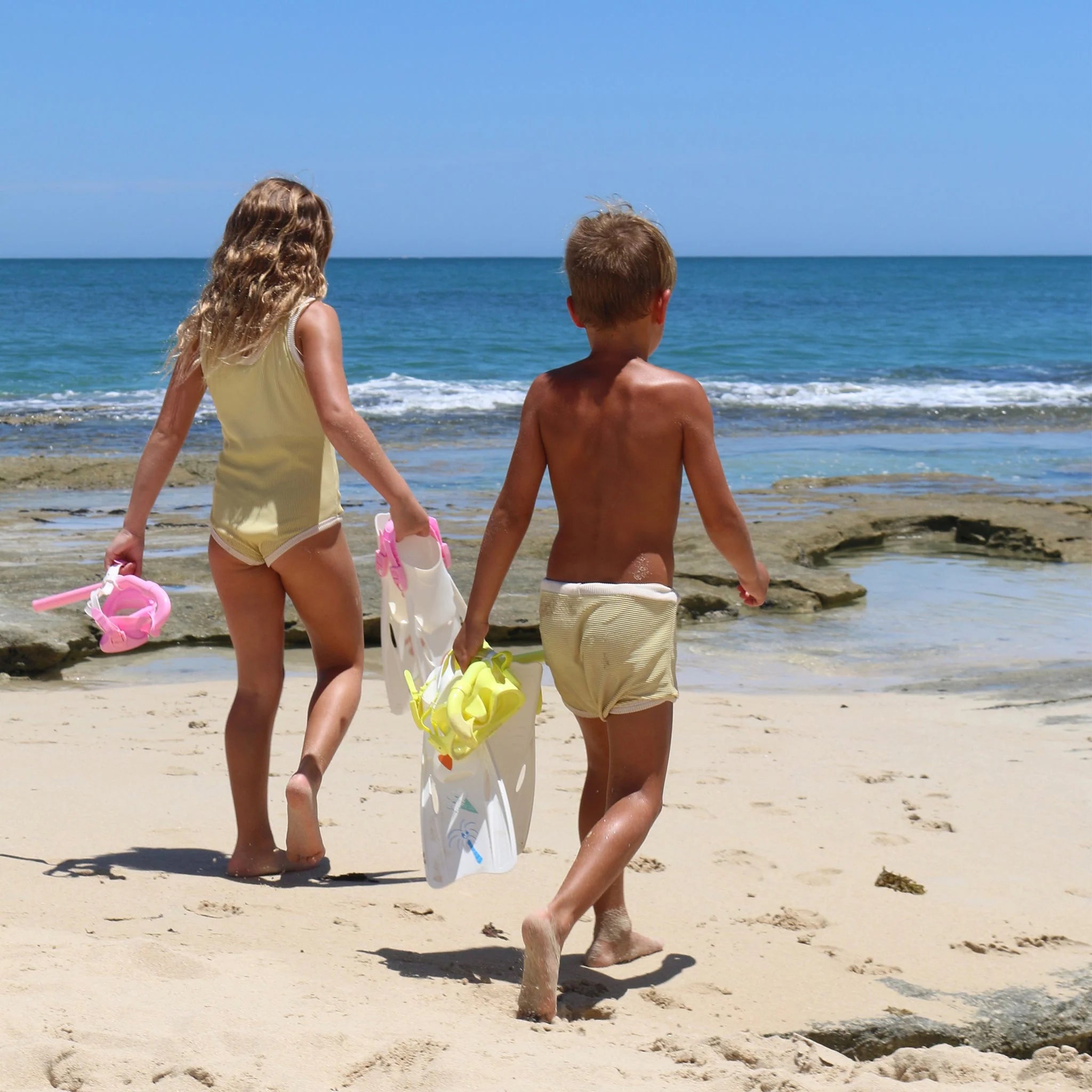 Two children in swimwear, carrying the SUNNYLIFE Kids Snorkel Set Pink with an adjustable strap, are walking toward the ocean on a sandy beach. The day is sunny with a clear blue sky and calm sea. There are rocks visible along the shoreline. The children are seen from the back.