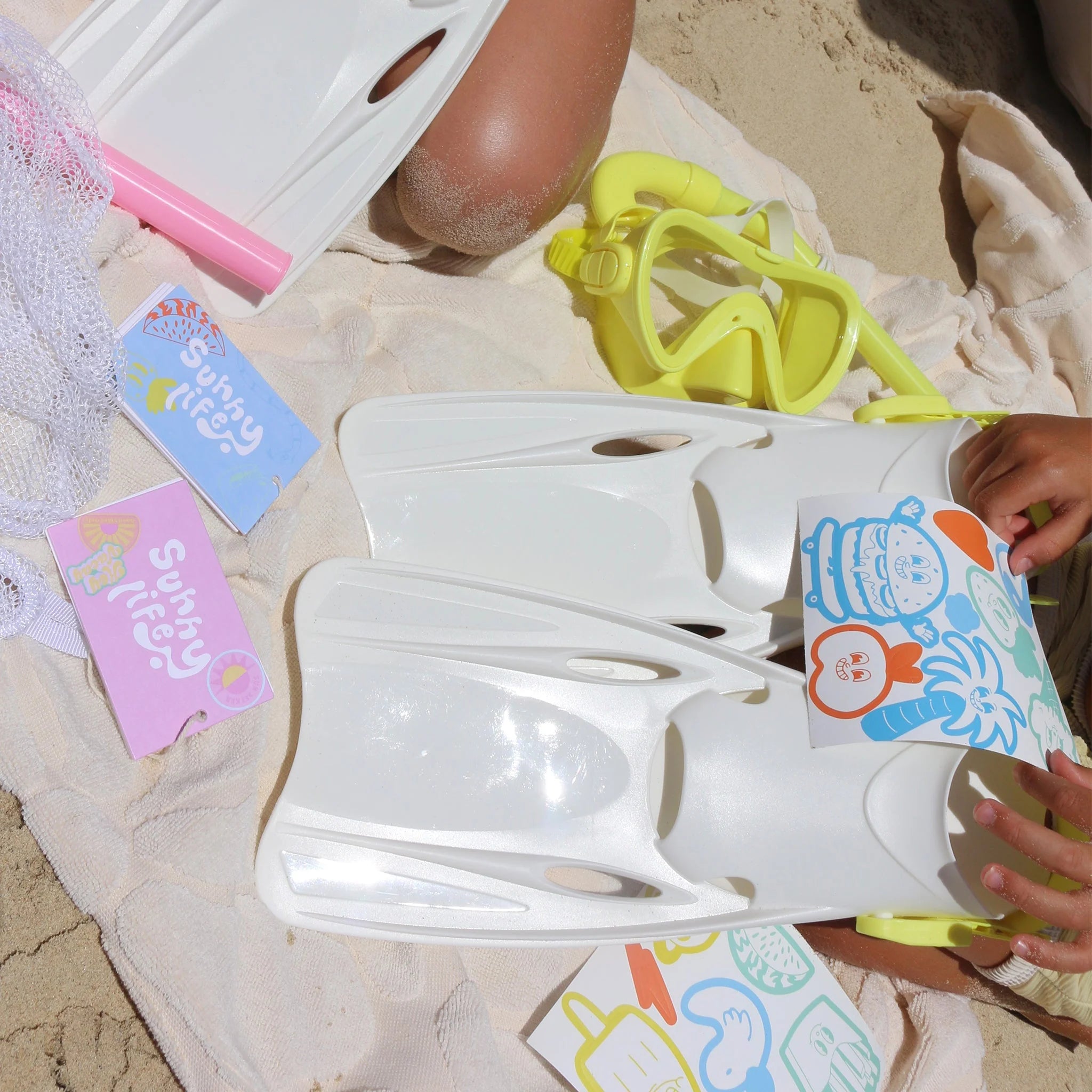 A beach scene featuring a child assembling the SUNNYLIFE Kids Snorkel Set Yellow, which includes yellow goggles, a snorkel, and white flippers, on a sandy surface with a towel. Stickers and postcards with colorful summer-themed designs, such as palm trees and beach balls, are scattered nearby.