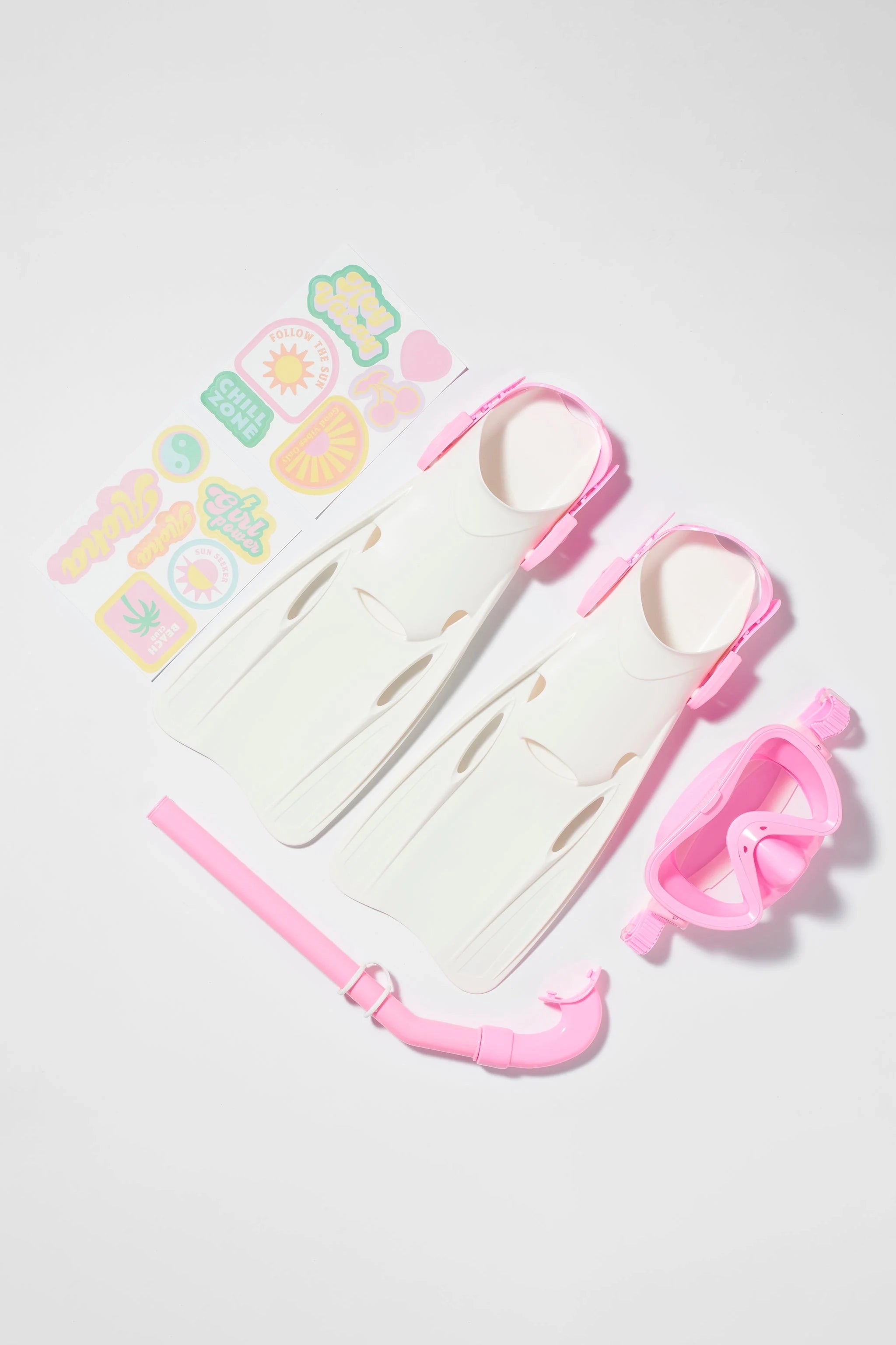 A SUNNYLIFE Kids Snorkel Set Pink, which features white and pink flippers, a snorkel mask with an adjustable strap, and a snorkel tube, is arranged neatly on a white background. Next to the snorkel set is a sheet of colorful customization stickers with various designs.