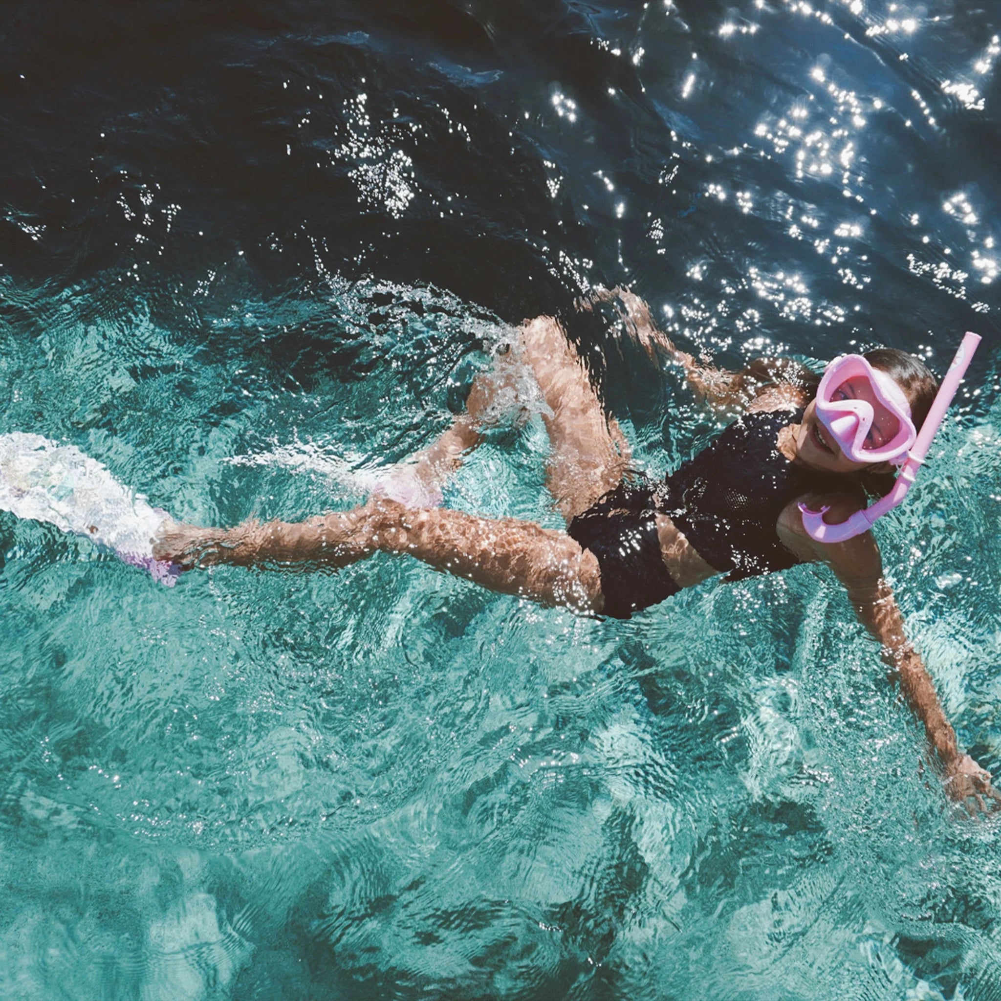 A person wearing a black swimsuit, a Kids Snorkel Set Pink by SUNNYLIFE with an adjustable strap, and swim fins floats on the surface of clear, sparkling water, surrounded by sunlight reflections. Their arms and legs are outstretched, suggesting relaxation and enjoyment of the aquatic environment.