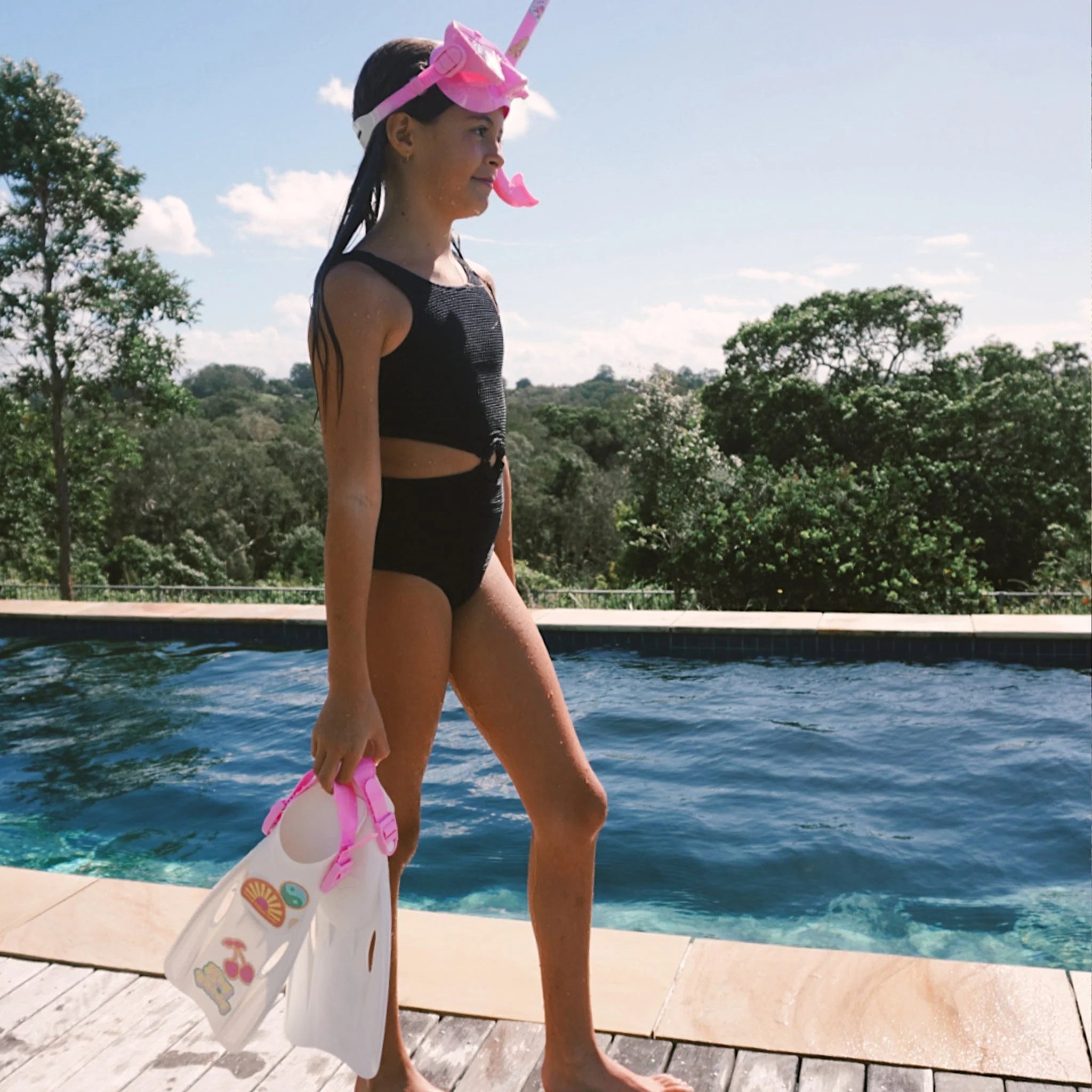 A young girl in a black swimsuit and the Kids Snorkel Set Pink by SUNNYLIFE stands by a pool, holding white flippers adorned with colorful customization stickers. The background shows lush green trees under a clear blue sky.