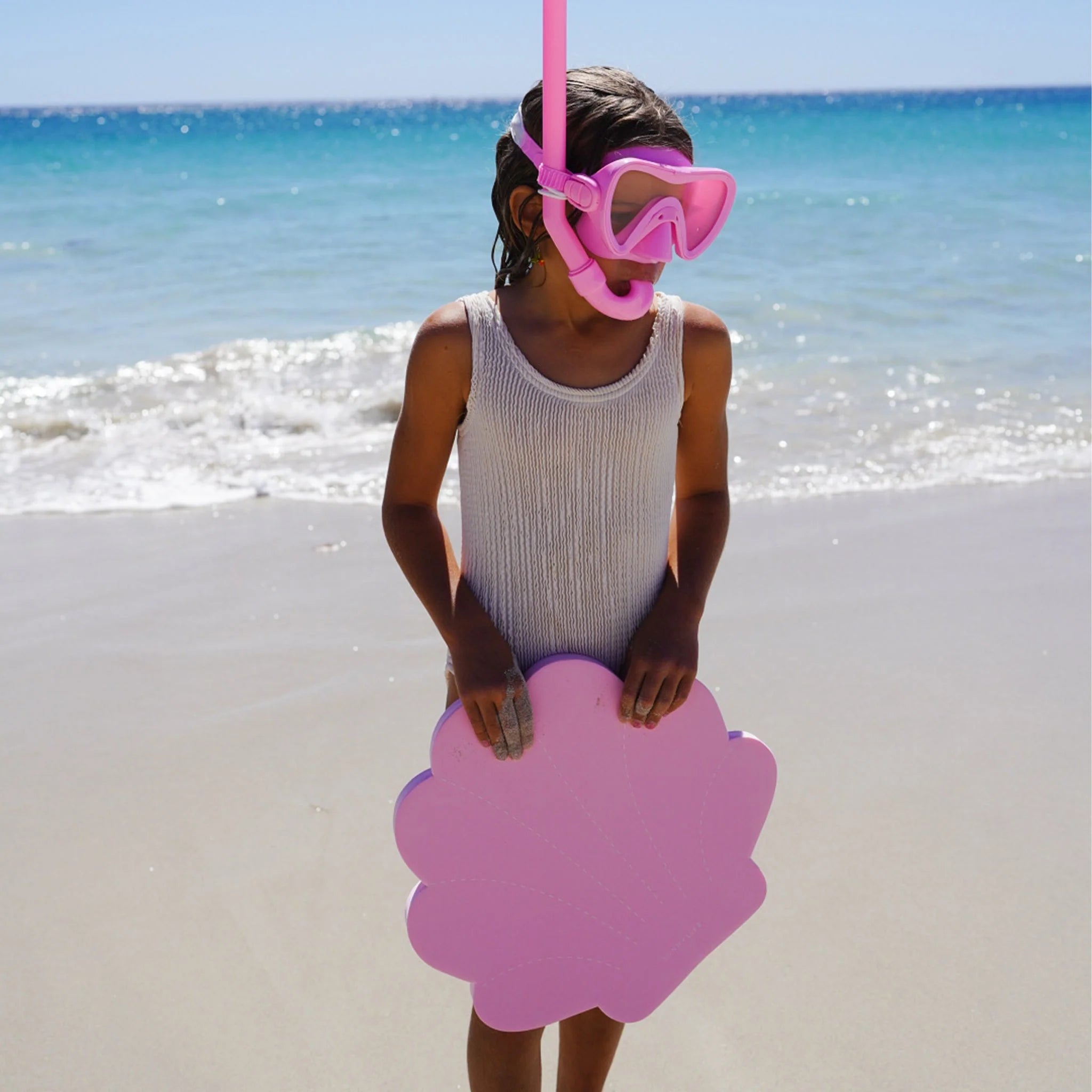 A child wearing a gray swimsuit and the SUNNYLIFE Kids Snorkel Set Pink stands on a beach, holding a pink shell-shaped float. The ocean waves gently roll onto the sandy shore in the background under a clear blue sky.