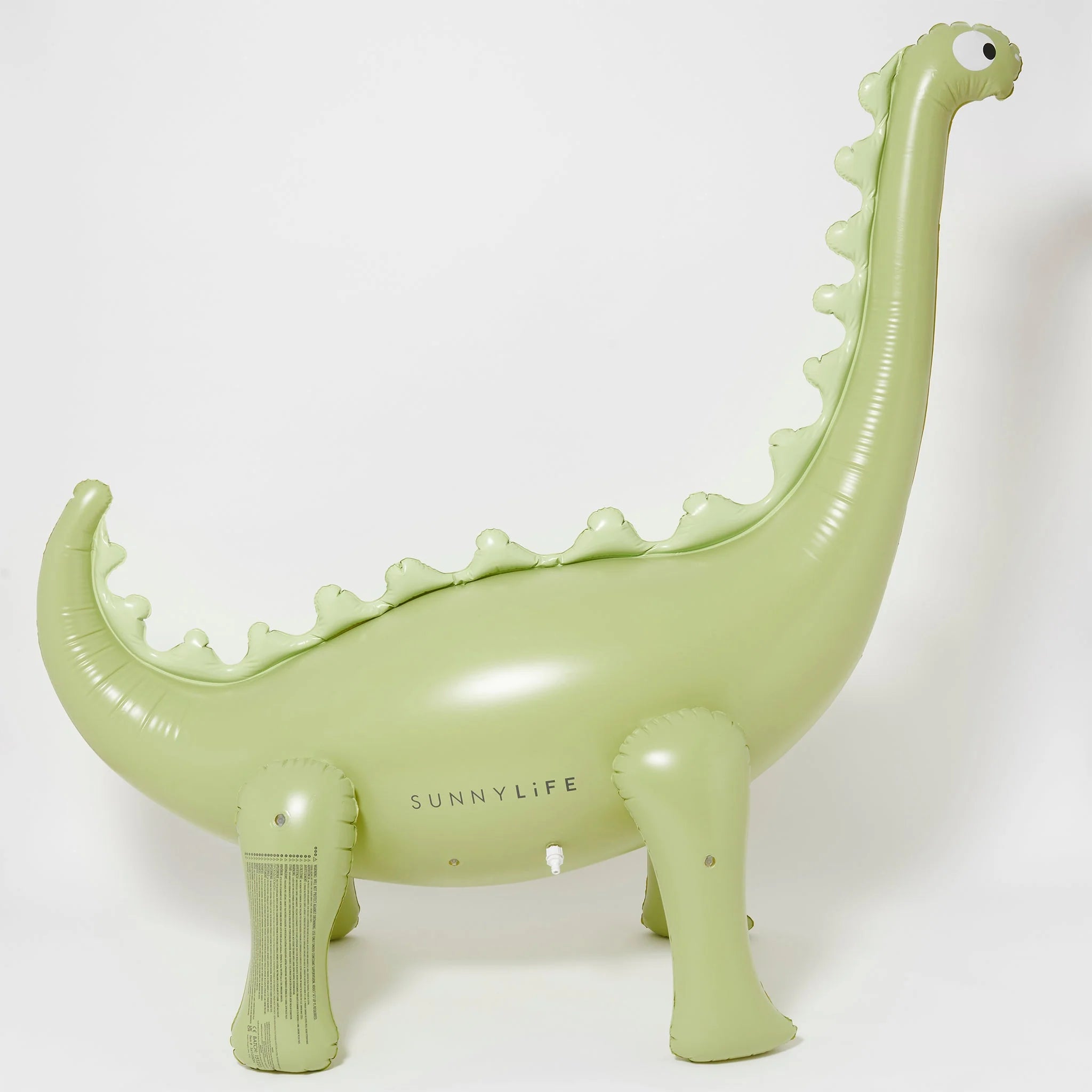 The Dino Giant Sprinkler is a light green inflatable pool float shaped like a dinosaur with four legs, a long neck, and a tail. Made from durable PVC, it features scalloped edges along its back and simple facial markings with nostrils and eyes. The brand name "SUNNYLIFE" is printed on its side, and it comes with a repair patch for convenience.