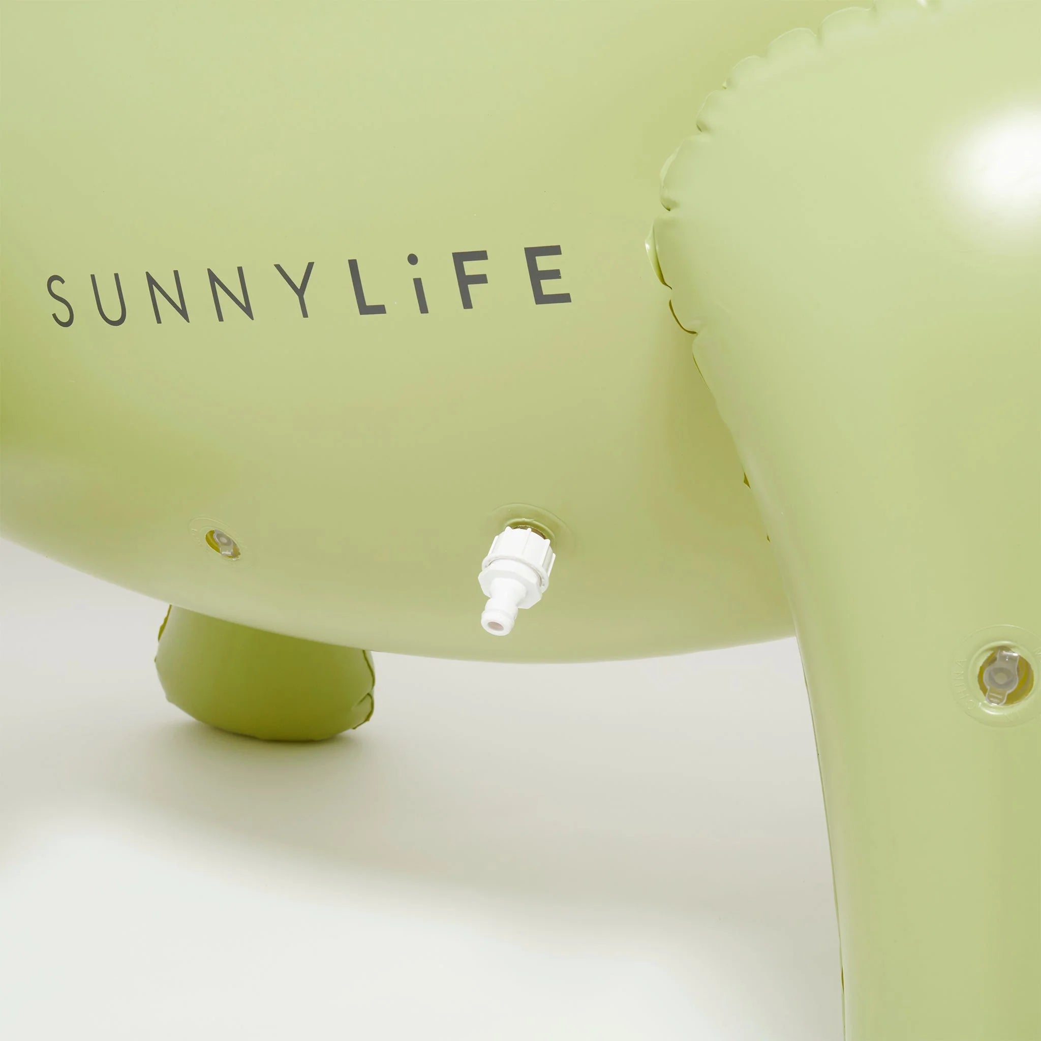 Close-up image of a light green inflatable object made from durable PVC, with the brand name "SUNNYLIFE" printed on it. A white valve is visible for inflating or deflating the item. The product is named "Dino Giant Sprinkler.