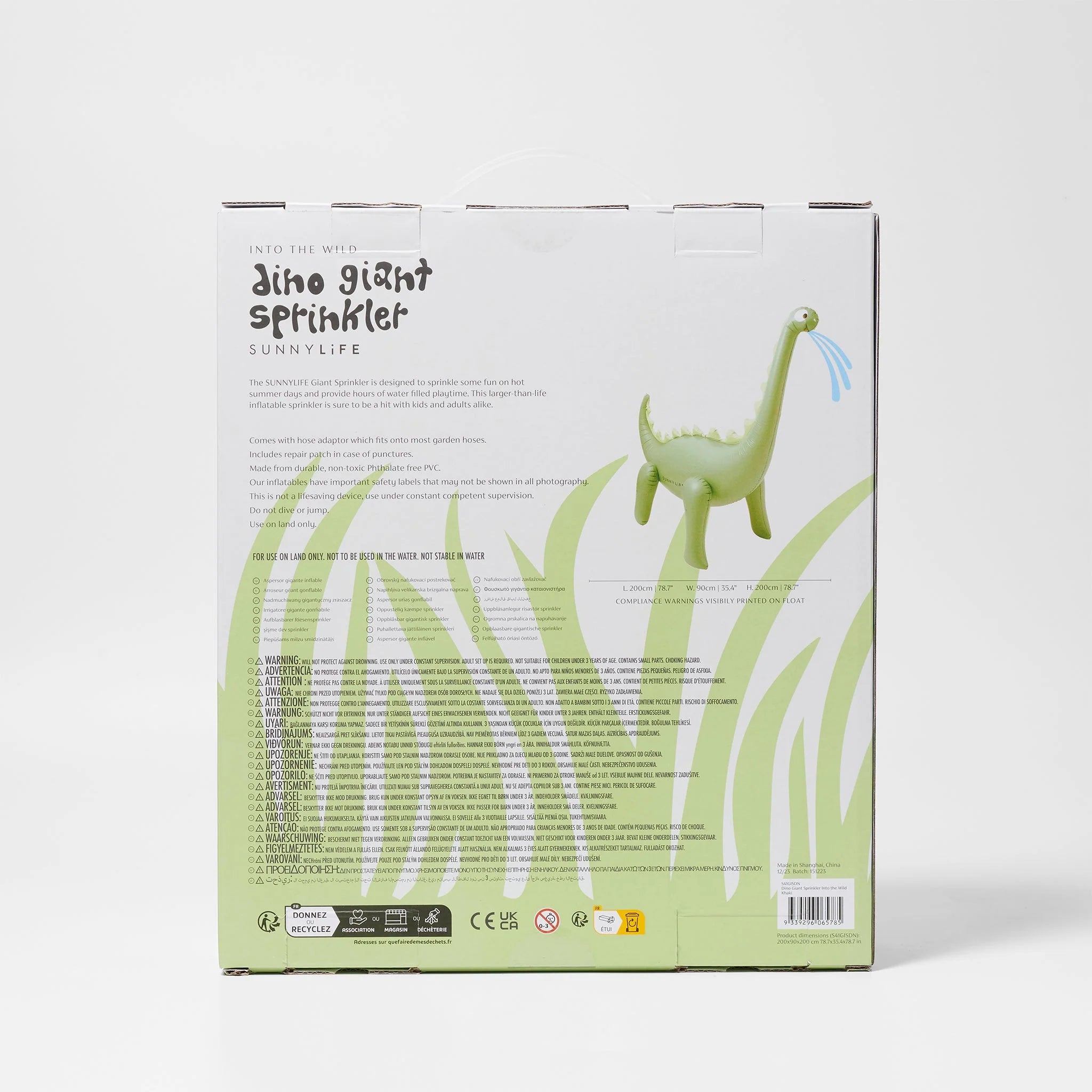 Back of a product box for the SUNNYLIFE Dino Giant Sprinkler. The box showcases an image of a green dinosaur-shaped sprinkler crafted from durable PVC material and lists various product details, user instructions, and safety warnings in multiple languages. A garden hose adaptor is also included. The background is adorned with tall grass illustrations.