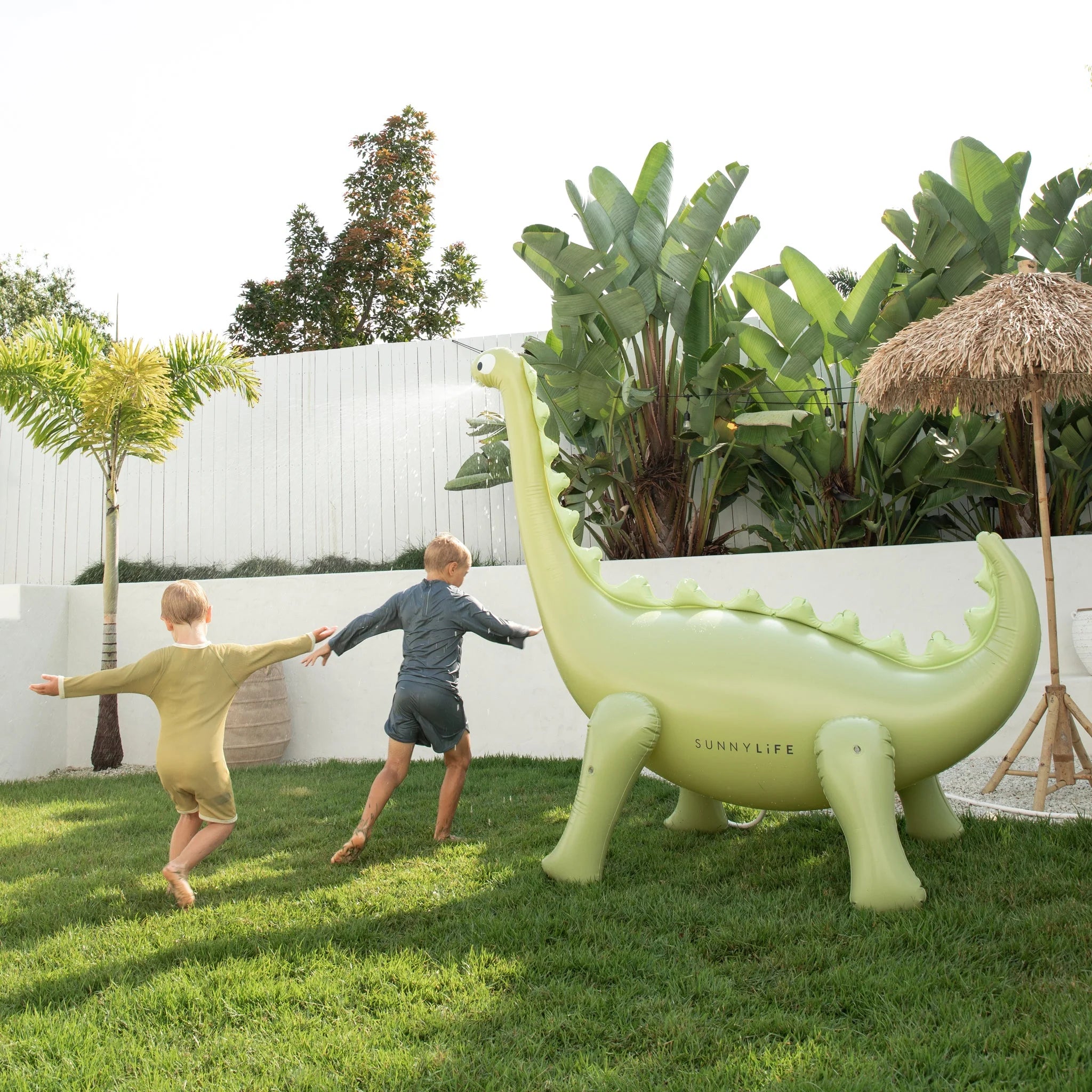 Two children play and hold hands, running in a grassy backyard near the Dino Giant Sprinkler, a large inflatable dinosaur made of durable PVC with "SUNNYLIFE" written on its side. The area has leafy plants, a thatched umbrella, and a white fence in the background.