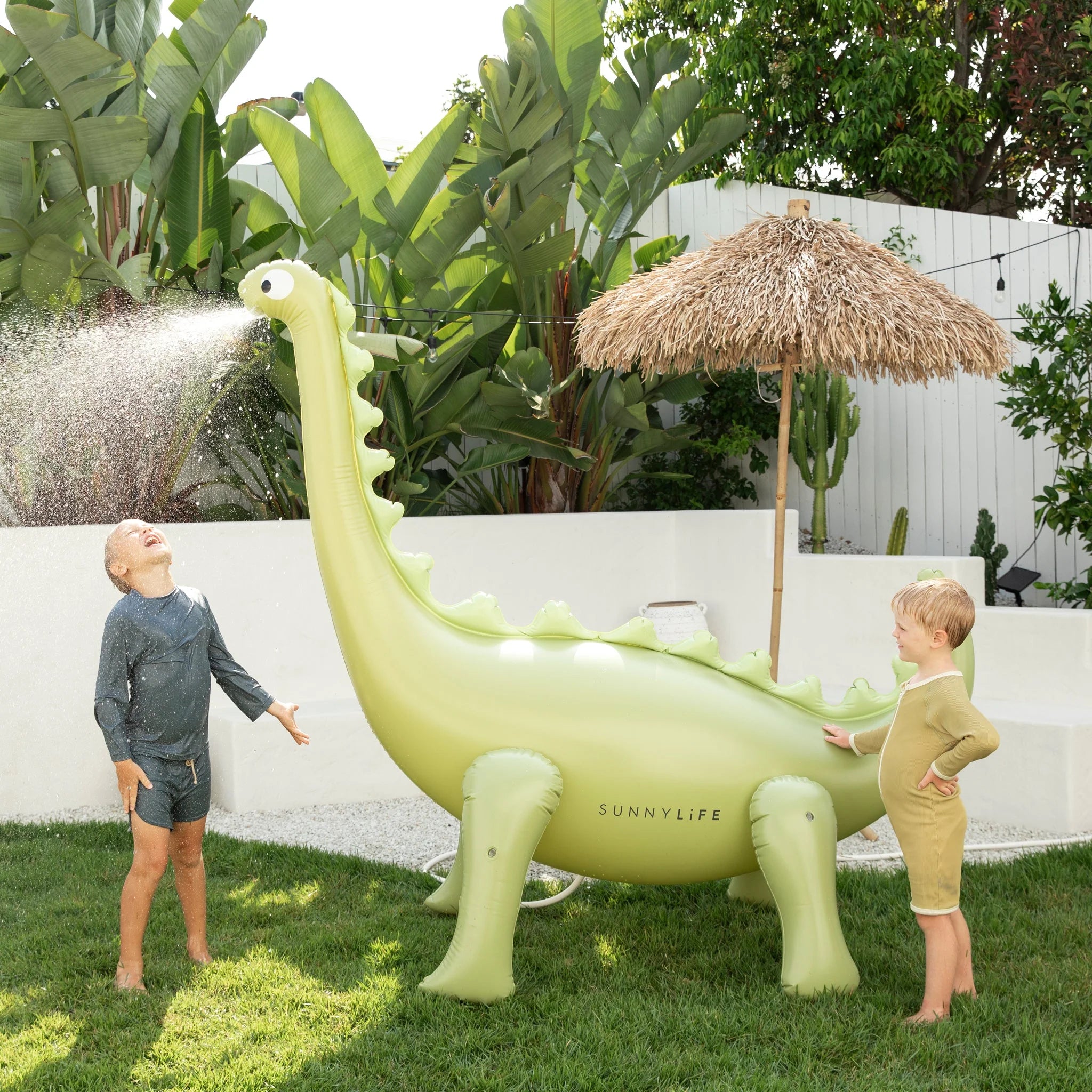 Two children are playing outside with the Dino Giant Sprinkler by SUNNYLIFE, a large green inflatable dinosaur crafted from durable PVC. One child stands under the dinosaur’s water spout, laughing as water sprays on them from the garden hose adaptor. The other child looks on, smiling. Lush greenery and a thatched umbrella provide the perfect backdrop.