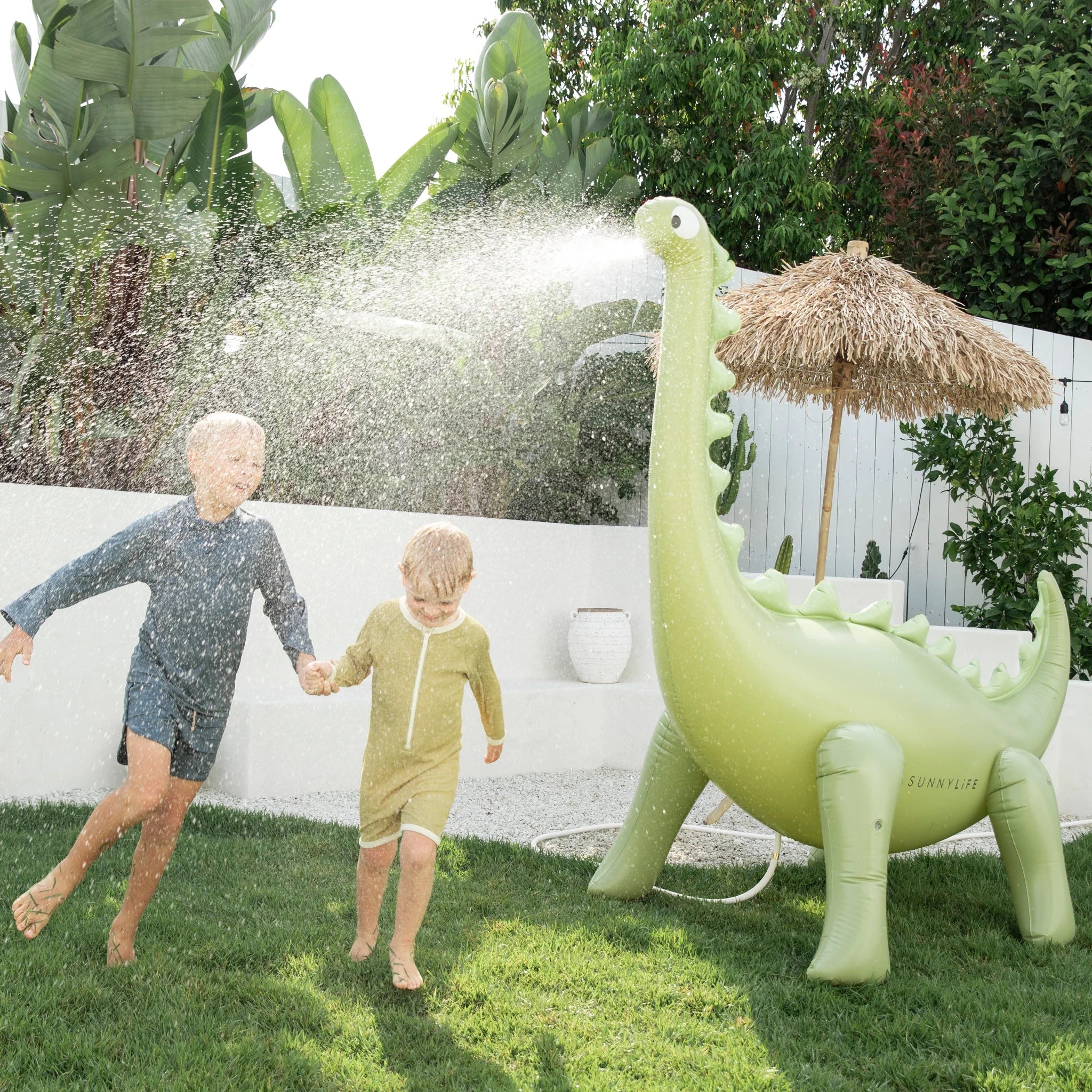 Two children, dressed in swimwear, run and play in a backyard with green grass. They are holding hands while being sprayed with water from the SUNNYLIFE Dino Giant Sprinkler made of durable PVC. Lush green plants and a white fence with a straw umbrella are in the background.