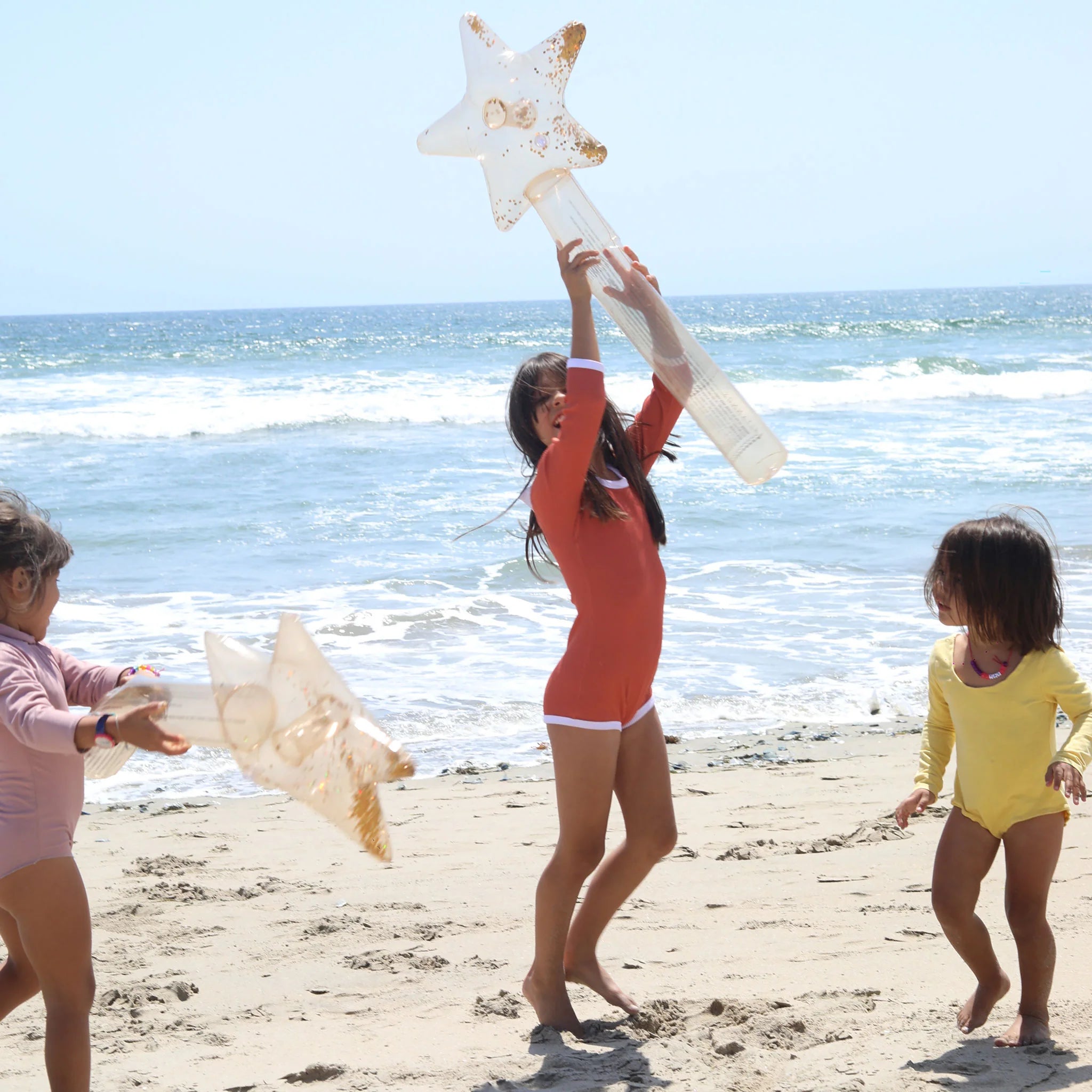 Three children, wearing brightly colored swimsuits, are playing with SUNNYLIFE Inflatable Star Wands (Set of 2) made from phthalate-free PVC on a sandy beach. The ocean waves are visible in the background under a clear, sunny sky. The middle child is in mid-spin, holding the wand above their head.