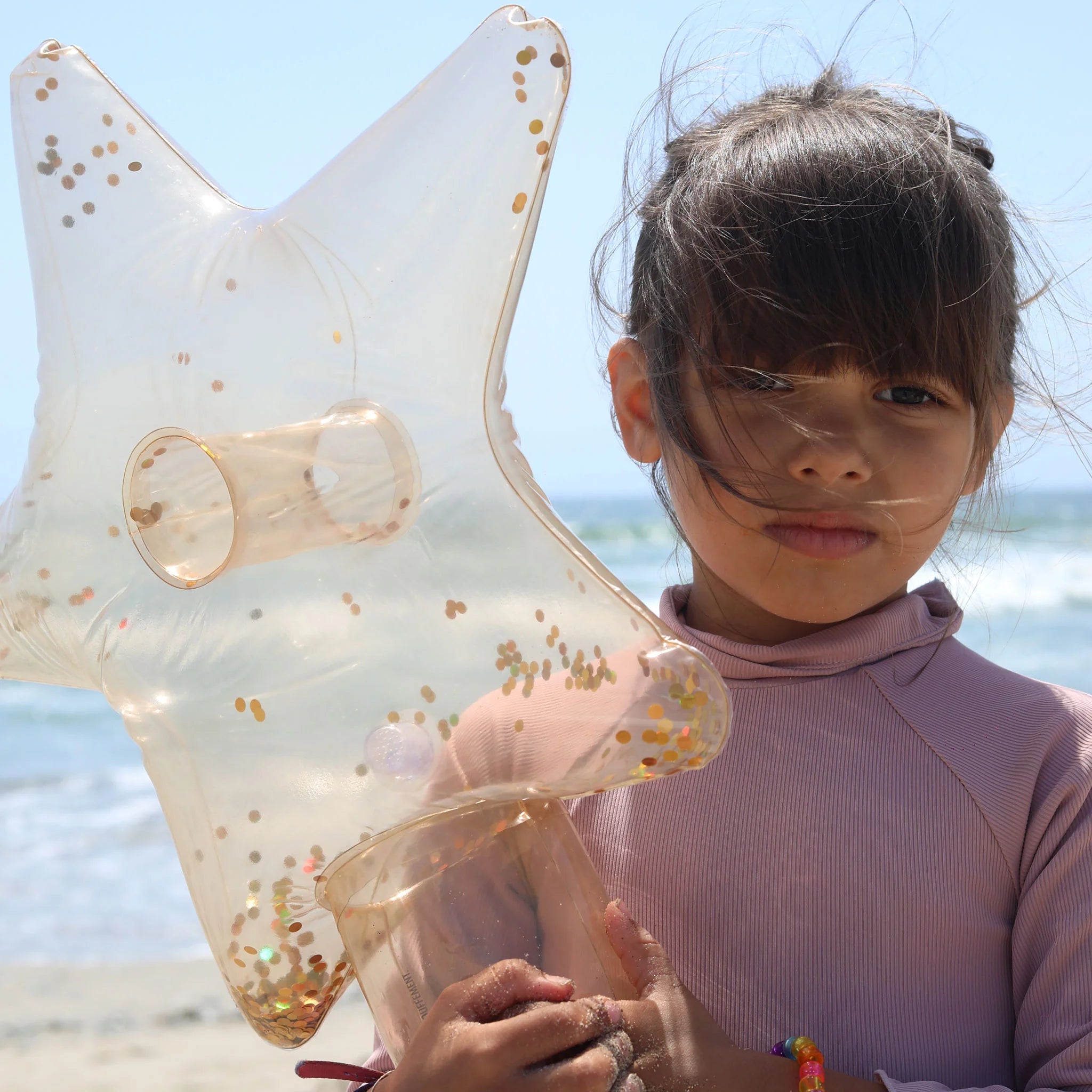 A young girl stands on a beach holding a transparent, star-shaped inflatable toy filled with gold glitter, crafted from Phthalate free PVC. She has long brown hair with bangs and wears a pink long-sleeve shirt. The ocean waves and blue sky are visible in the background. The toy she is holding is part of SUNNYLIFE's Inflatable Star Wands (Set of 2).