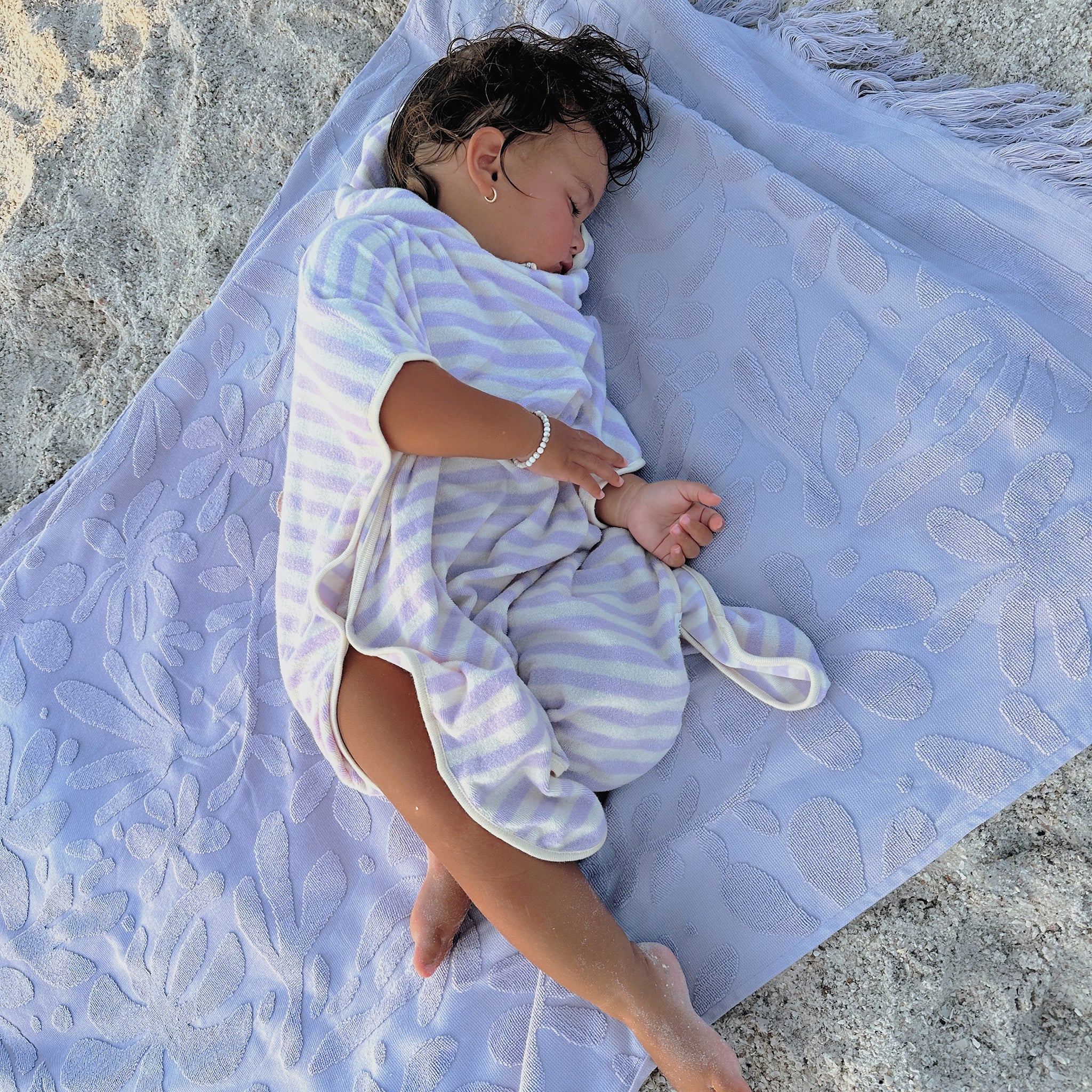 A young child, wrapped in a lavender and white striped blanket made of soft French Terry, sleeps peacefully on a SUNNYLIFE Kids Character Hooded Towel Princess Swan/Soft Lilac laid out on the sand. The child has messy dark hair and is wearing a small bracelet from their favorite kids clothing brand.