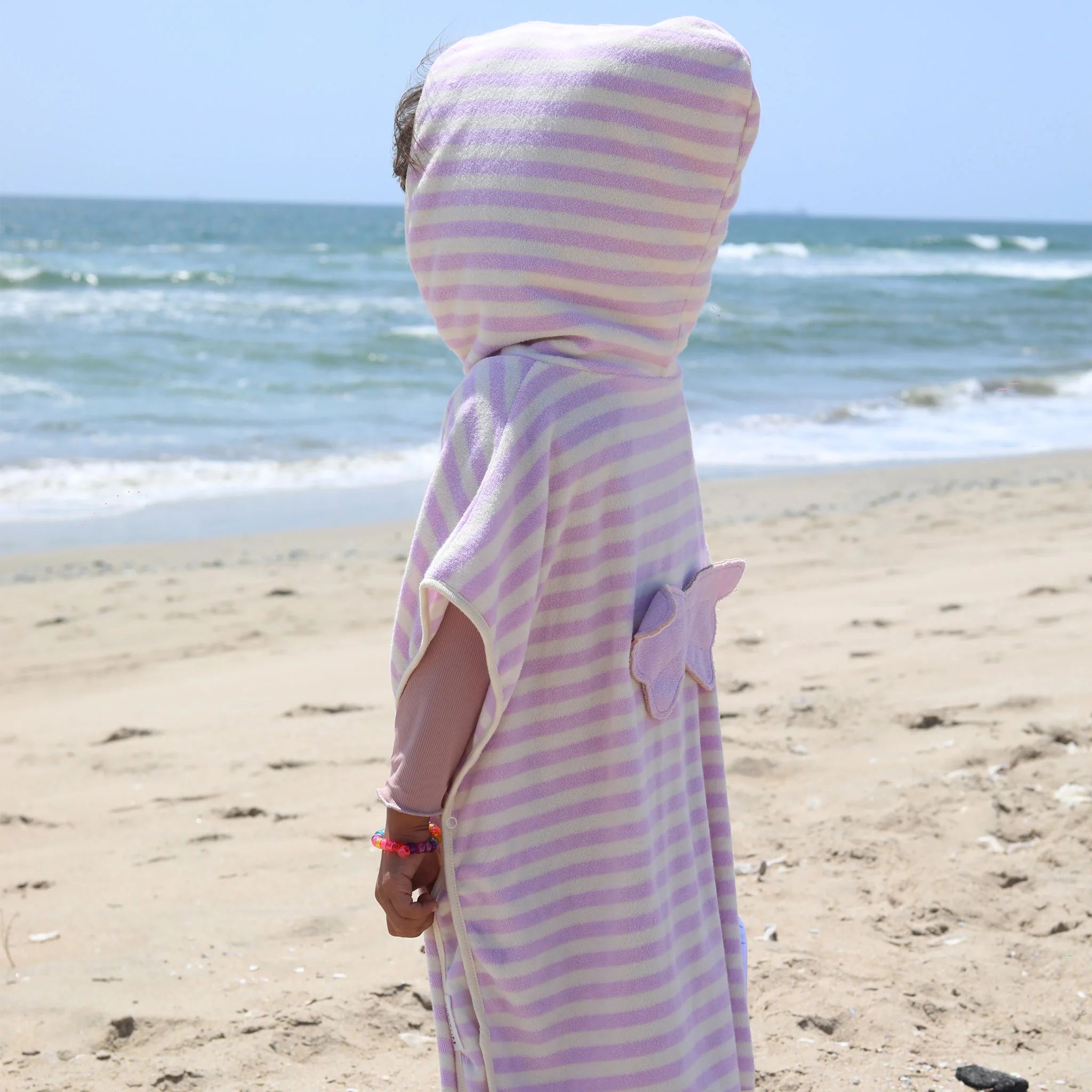 A child wrapped in a SUNNYLIFE Kids Character Hooded Towel in the Princess Swan/Soft Lilac design stands at the edge of the beach, facing the ocean waves. The soft French Terry towel features a starfish-shaped patch with a 3D character element on the back. With a bracelet on their wrist, the child appears to be gazing out towards the sea.