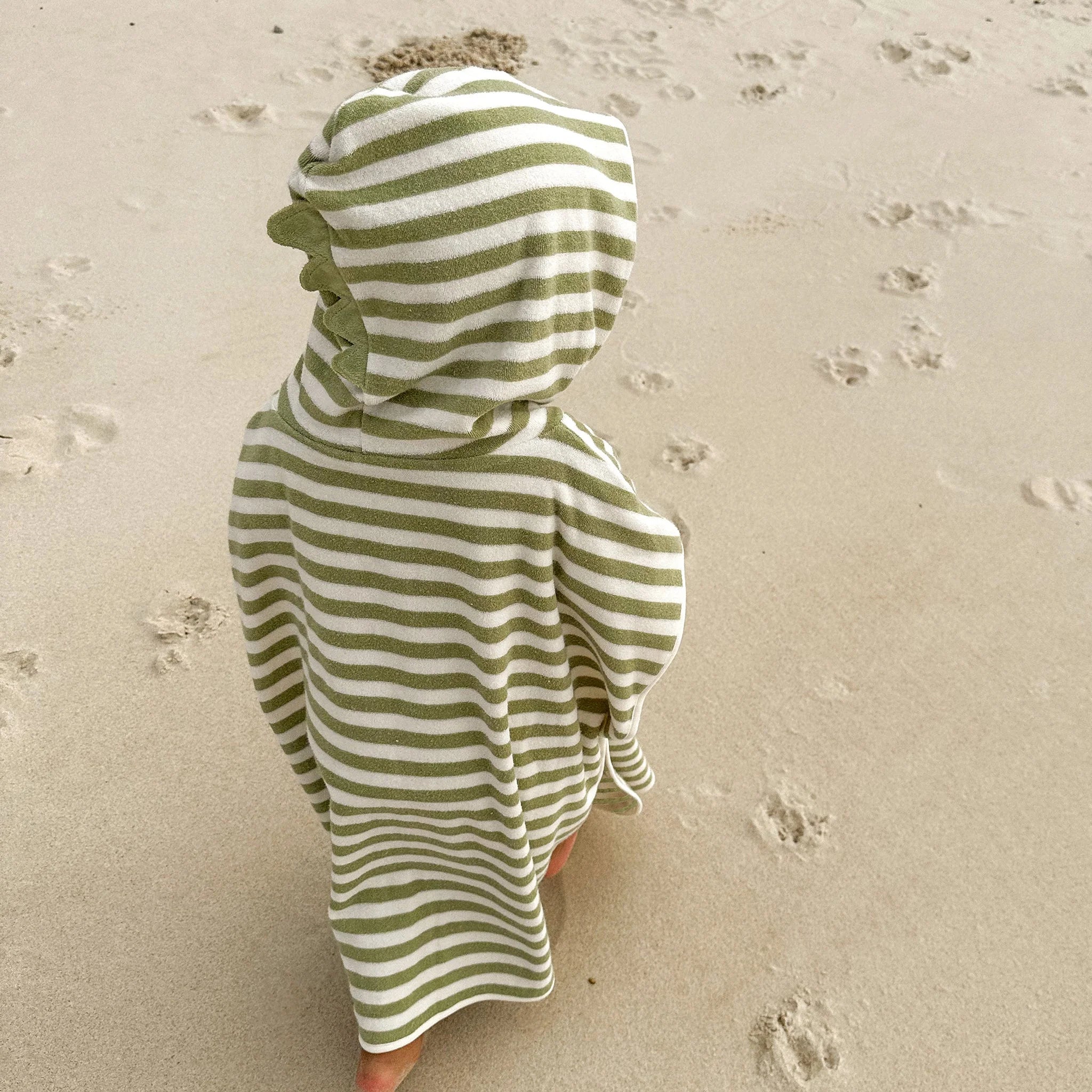 A child wrapped in a SUNNYLIFE Kids Character Hooded Towel Into the Wild/Khaki stands on a sandy beach. The child is facing away from the camera, seemingly looking at the ground. There are footprints and paw prints adorning the sand, adding character to this charming kids product scene.