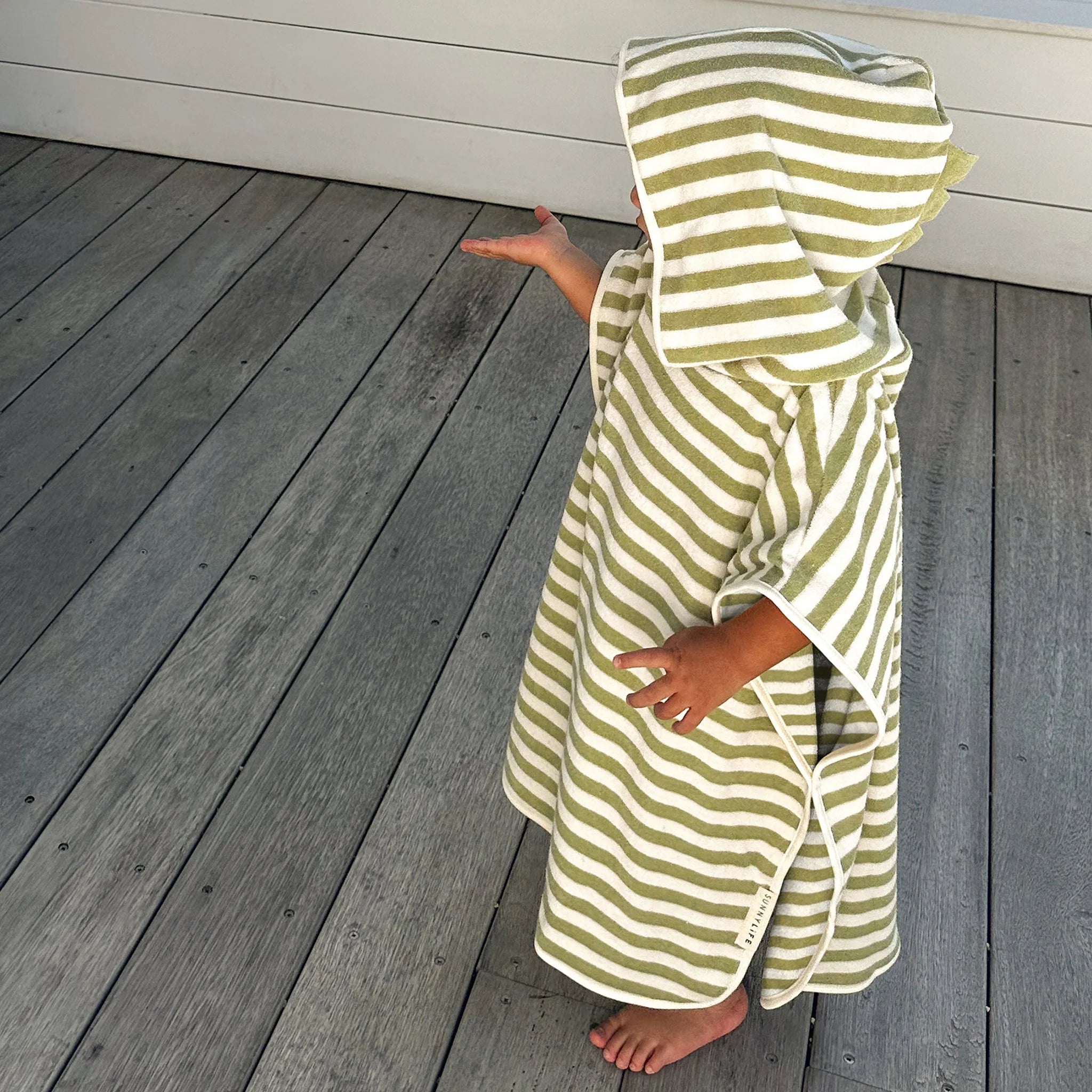 A small child, wrapped in a SUNNYLIFE Kids Character Hooded Towel in the Into the Wild/Khaki design made of soft French Terry, stands on a wooden deck. The kid extends their right arm while gazing in that direction, the hood covering part of their head and face. This charming scene could be straight out of a 3D character element design for a kids product.