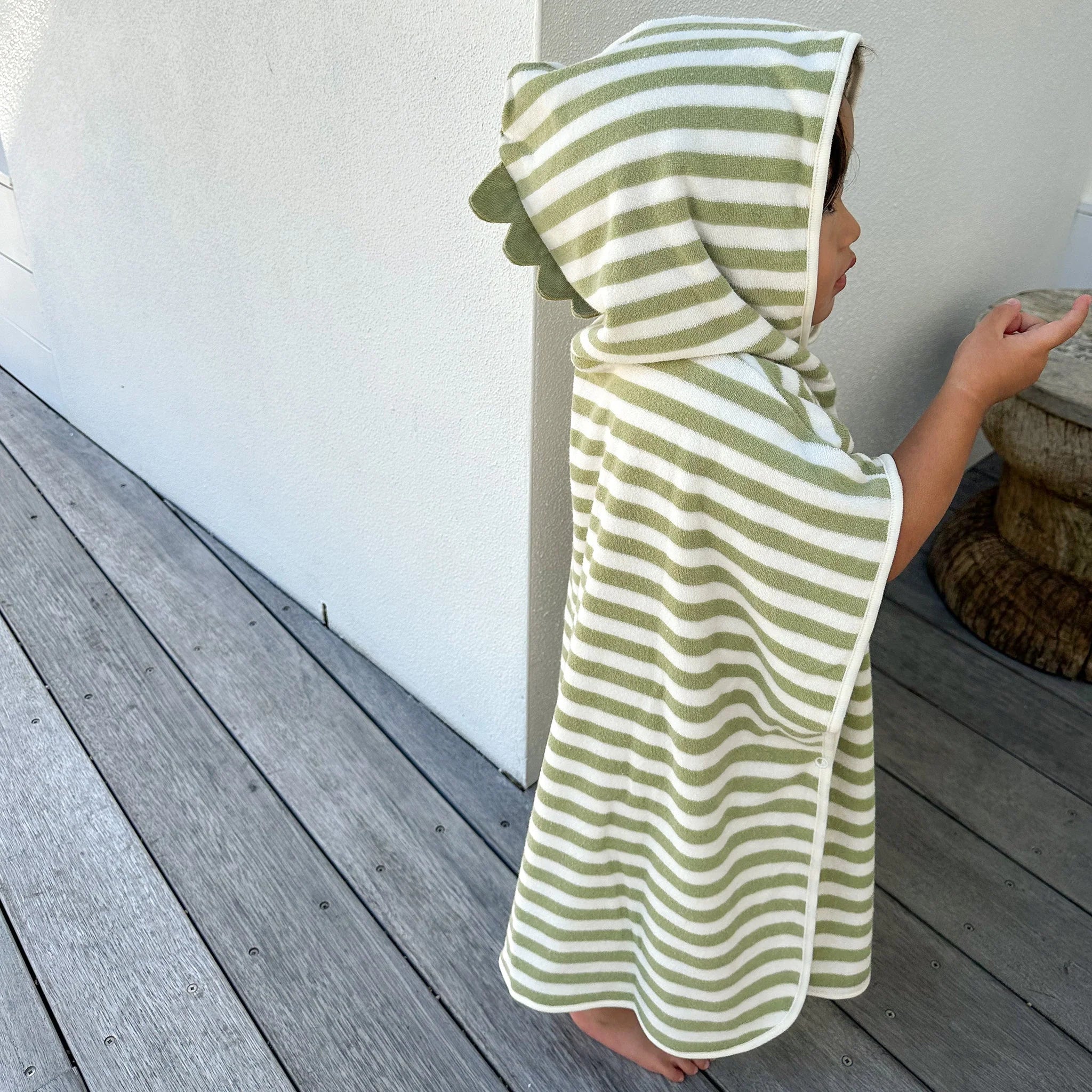 A child stands on a wooden deck wearing the SUNNYLIFE Kids Character Hooded Towel Into the Wild/Khaki, made of soft French Terry with green and khaki stripes. The hood features small green triangular flaps resembling dinosaur scales. The child is pointing at something off-frame, adding a playful 3D character element to this charming kids' product.