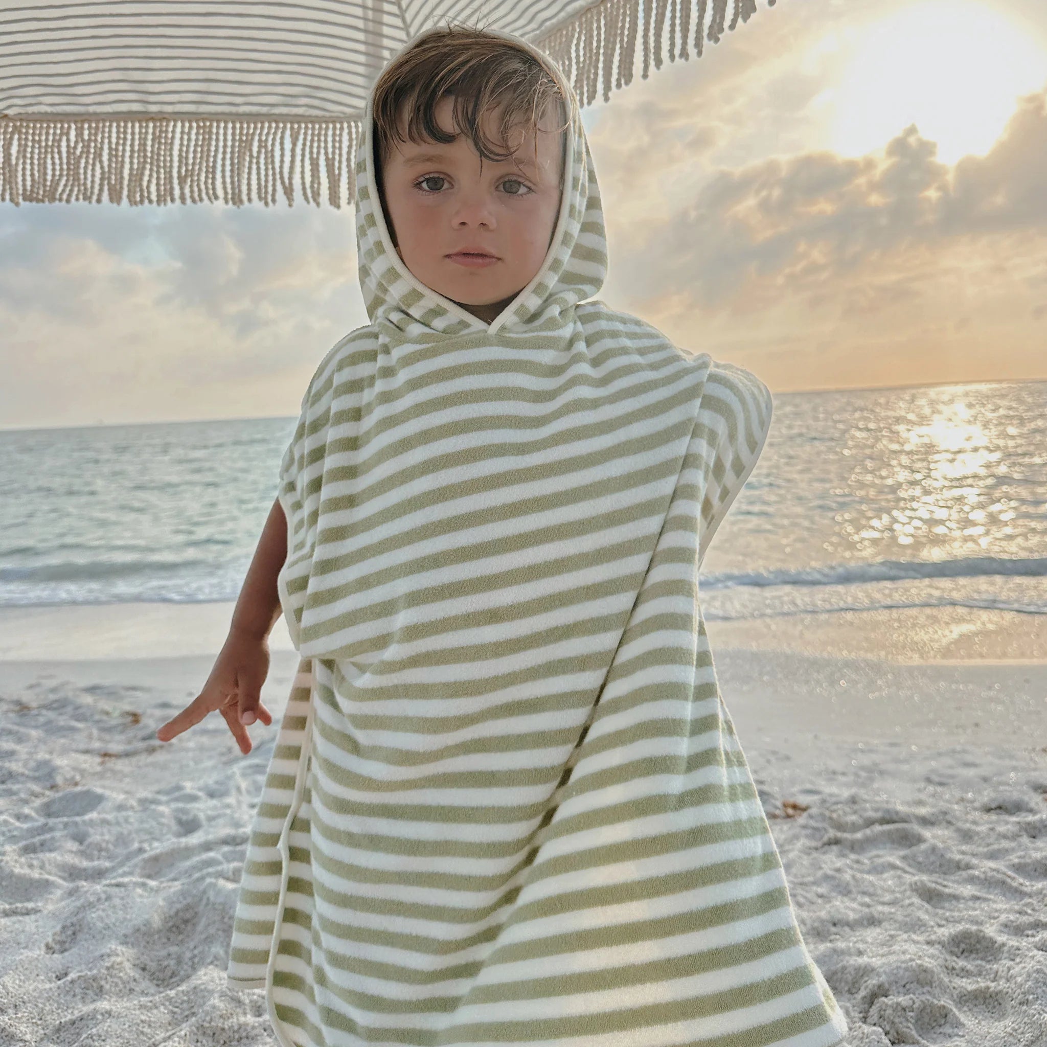 A child stands on a sandy beach wrapped in the SUNNYLIFE Kids Character Hooded Towel in the Into the Wild/Khaki design. An umbrella with fringe provides shade, while the sun sets over a calm sea in the background, casting a warm glow on the scene. The child looks directly at the camera with a serene expression.