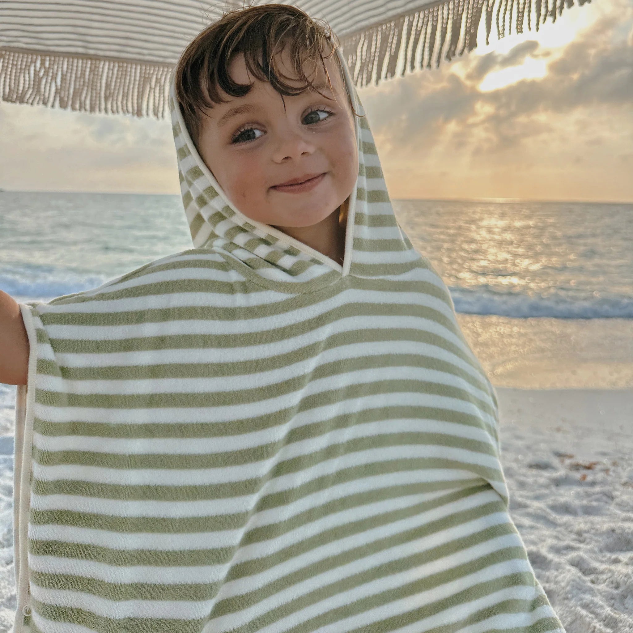 A child wearing a khaki "Kids Character Hooded Towel Into the Wild" by SUNNYLIFE smiles while standing under a fringed beach umbrella. The background features a sandy beach, gentle waves, and a sunset over the ocean, completing this charming kids product scene.