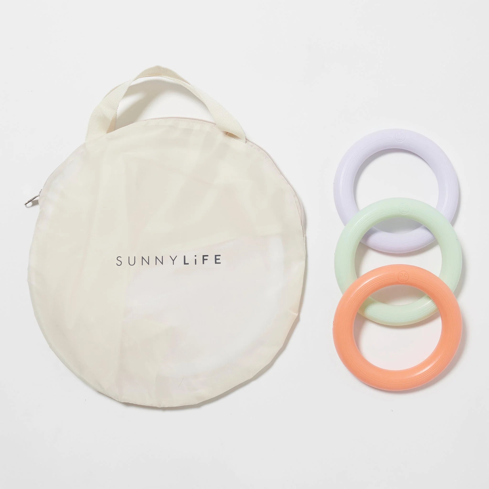 A round, cream-colored fabric storage bag with a handle and the word "SUNNYLIFE" printed on it sits beside three circular, pastel-hued rings in lavender, mint green, and peach made from durable non-toxic PVC. These items are part of the Baby Playmat with Shade by SUNNYLIFE and are arranged on a white background.