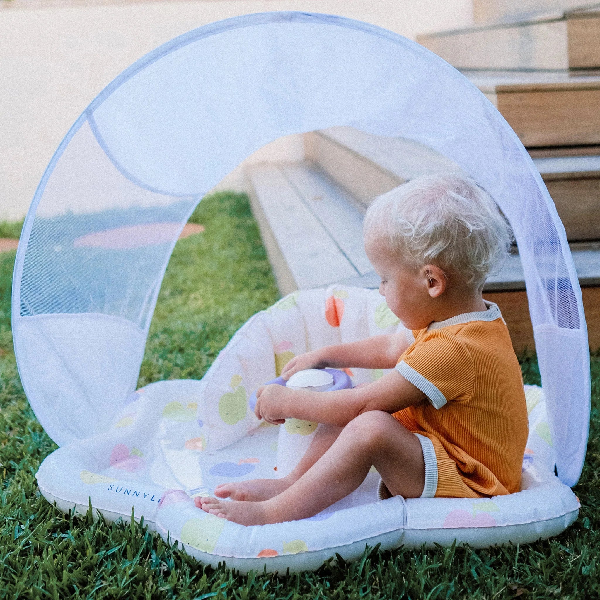 A young child with blonde hair, wearing an orange shirt, sits in a shaded *Baby Playmat with Shade* by SUNNYLIFE on a grassy lawn. The play area, made from durable non-toxic PVC, features a UPF40 canopy and colorful designs. Wooden steps are visible in the background as the child appears to be playing with a toy.