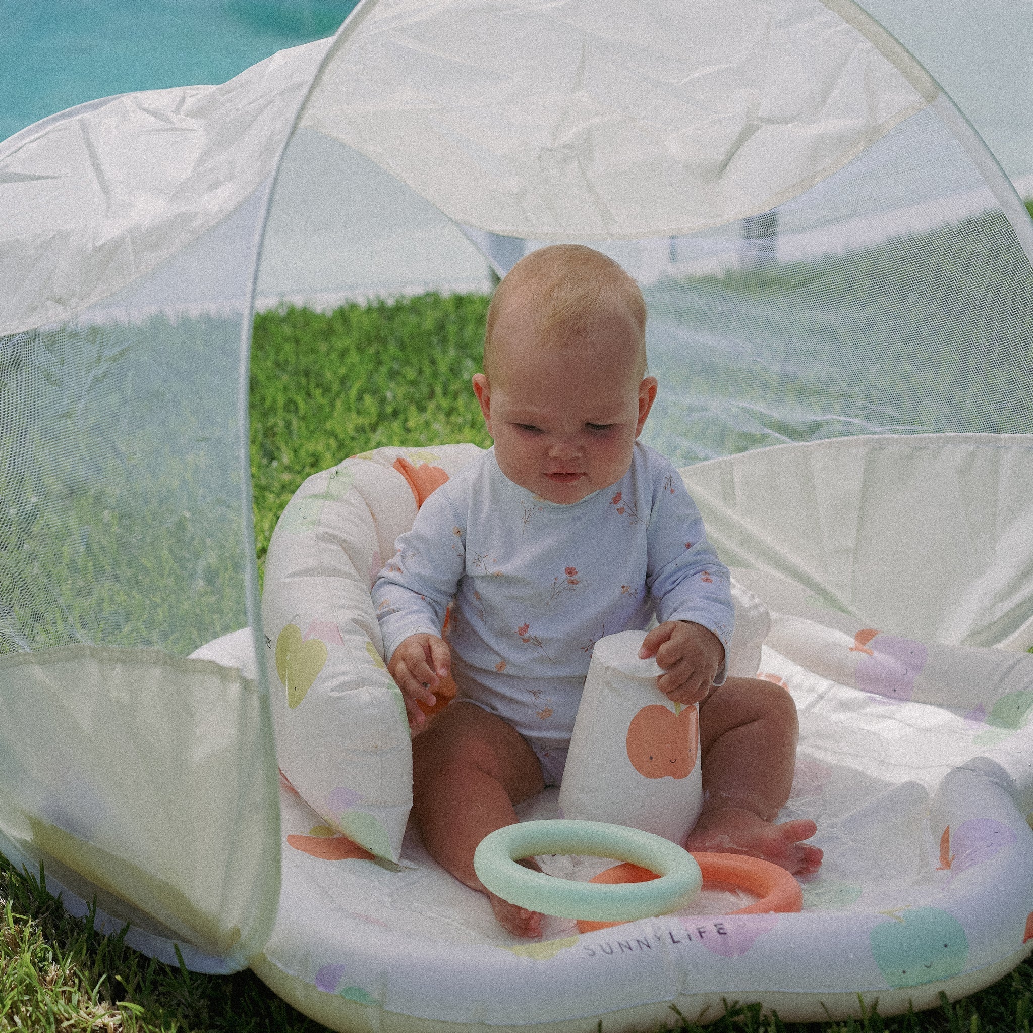 A baby sits on the SUNNYLIFE Baby Playmat with Shade, enjoying the colorful cushioned mat inside a small outdoor tent featuring a UPF40 canopy. The baby holds a white cup and is surrounded by various toys, including a light green ring. Positioned on the grass near a pool, this setup fosters hand-eye coordination. The baby is dressed in a long-sleeved patterned onesie.