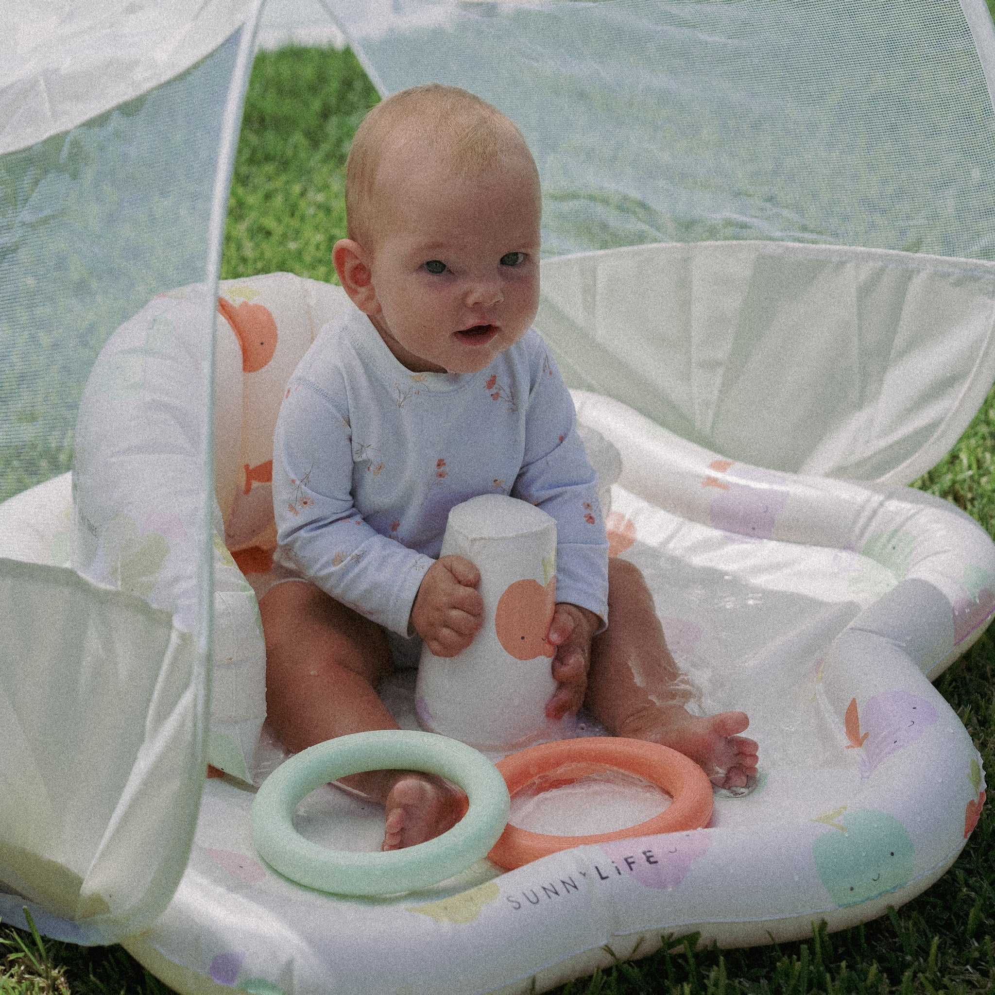 A baby sits inside the SUNNYLIFE Baby Playmat with Shade on grass, wearing a light-colored outfit. Beneath the UPF40 canopy, the baby holds a toy cup with colorful rings nearby on the cushioned play mat. The pastel-patterned space supports hand-eye coordination with its engaging toys.