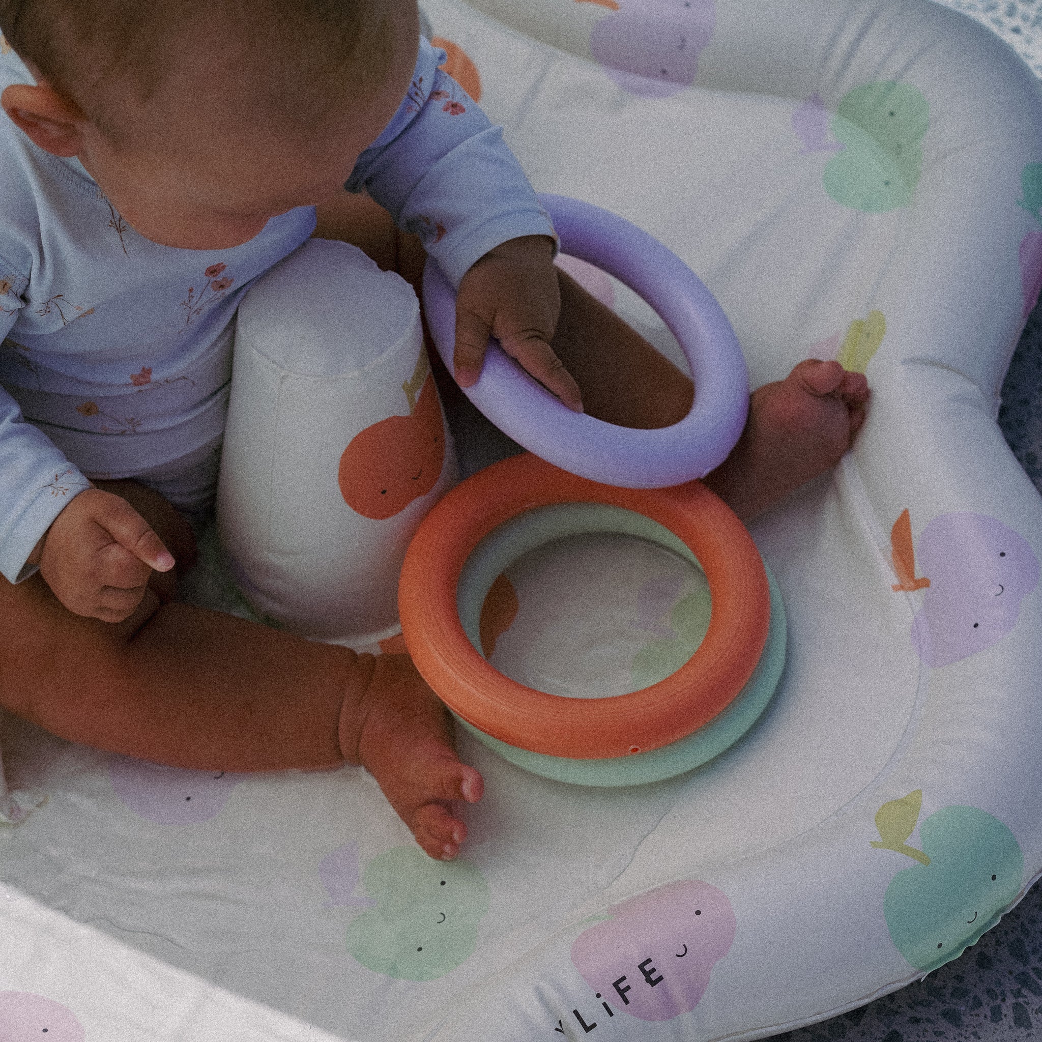 A baby sits joyfully in a SUNNYLIFE Baby Playmat with Shade, made from durable non-toxic PVC and decorated with pastel-colored balloons. Wearing a light-colored outfit adorned with small designs, the baby holds a purple ring toy in one hand, enhancing hand-eye coordination, while other colorful rings lay on the playmat.