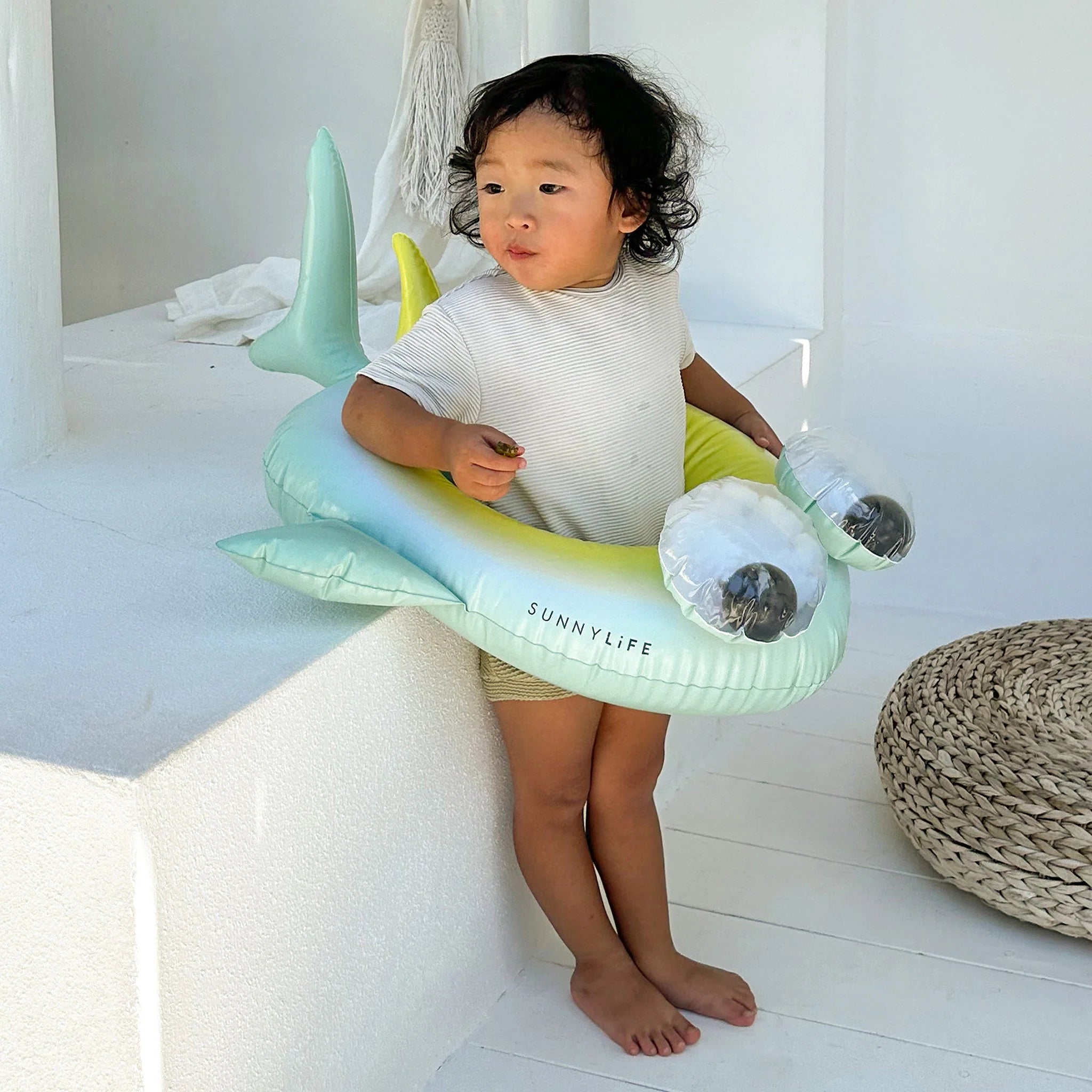 A toddler with curly hair stands indoors wearing a white and grey striped shirt and beige shorts. The child is holding the SUNNYLIFE Kids Tube Pool Ring Salty the Shark, crafted from durable PVC with white eyes and black pupils. The minimalistic setting includes white walls and a woven pouf in the background.