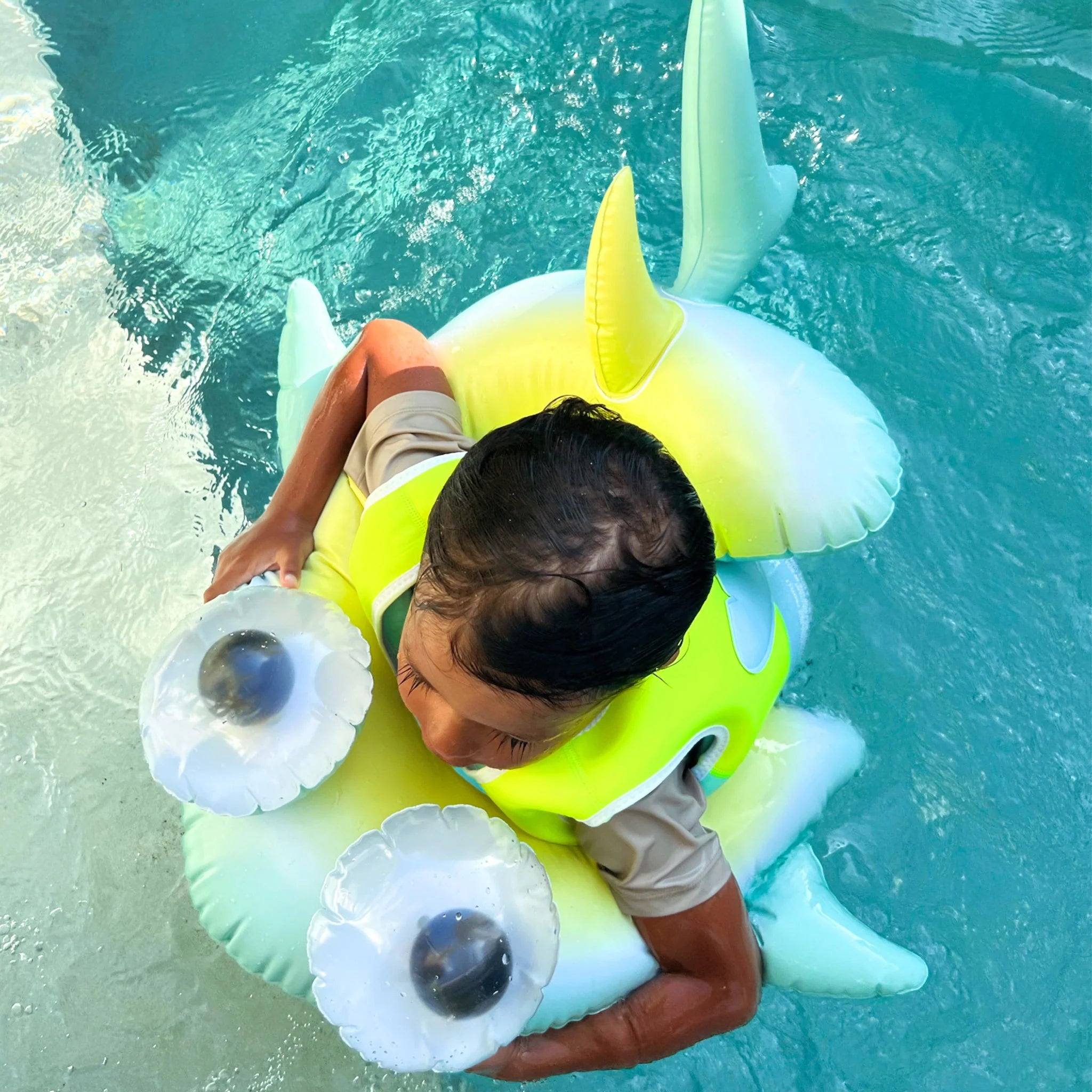 A child in a swimming pool is using the colorful Kids Tube Pool Ring Salty the Shark, produced by SUNNYLIFE, which is made from durable PVC. The float, featuring a combination of teal, yellow, and white colors, supports the child wearing a green life jacket. With wet hair and smiles all around, they enjoy the clear, slightly rippling water.