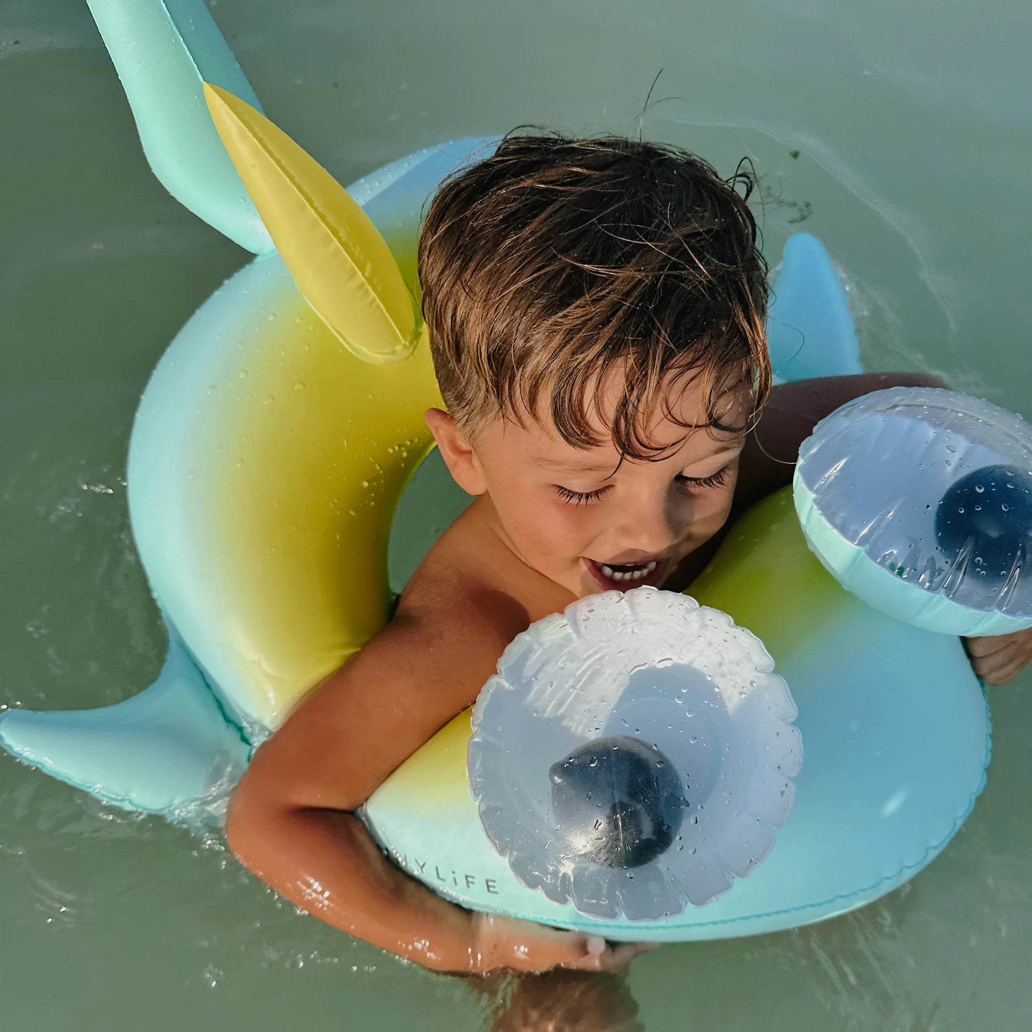 A young boy plays in shallow water while sitting in a blue and yellow inflatable sea animal float named "Salty the Shark," made of durable PVC by SUNNYLIFE. He holds onto the float and looks down at two transparent, round objects attached to it. The water around him is calm and clear.