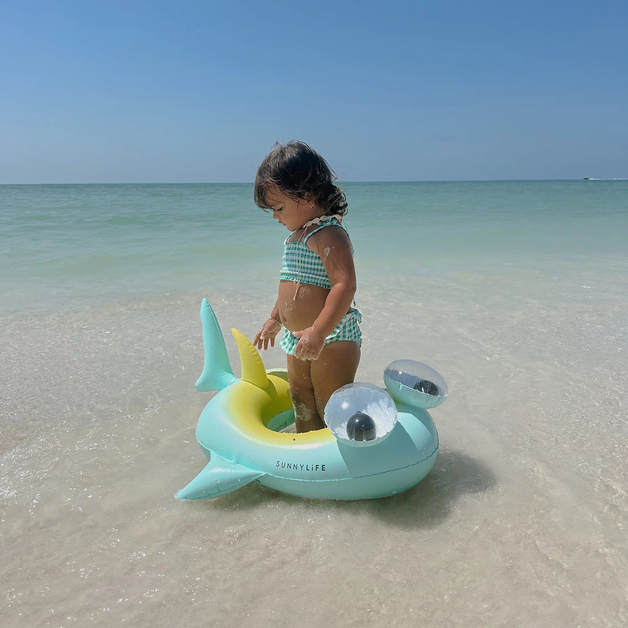 A young child stands in the vibrant Kids Tube Pool Ring Salty the Shark by SUNNYLIFE, made from durable PVC, on a sandy beach with gentle waves and a clear blue sky. The child is wearing a green and white striped swimsuit, looking down while enjoying the calm water.