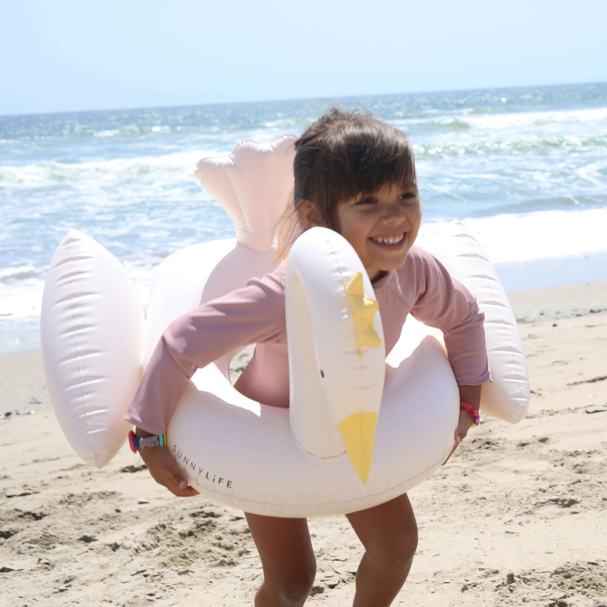 A young girl laughs joyfully while wearing a SUNNYLIFE Kids Tube Pool Ring Princess Swan around her waist at a sandy beach. The ocean waves are gently crashing in the background, and the sky is clear and blue.